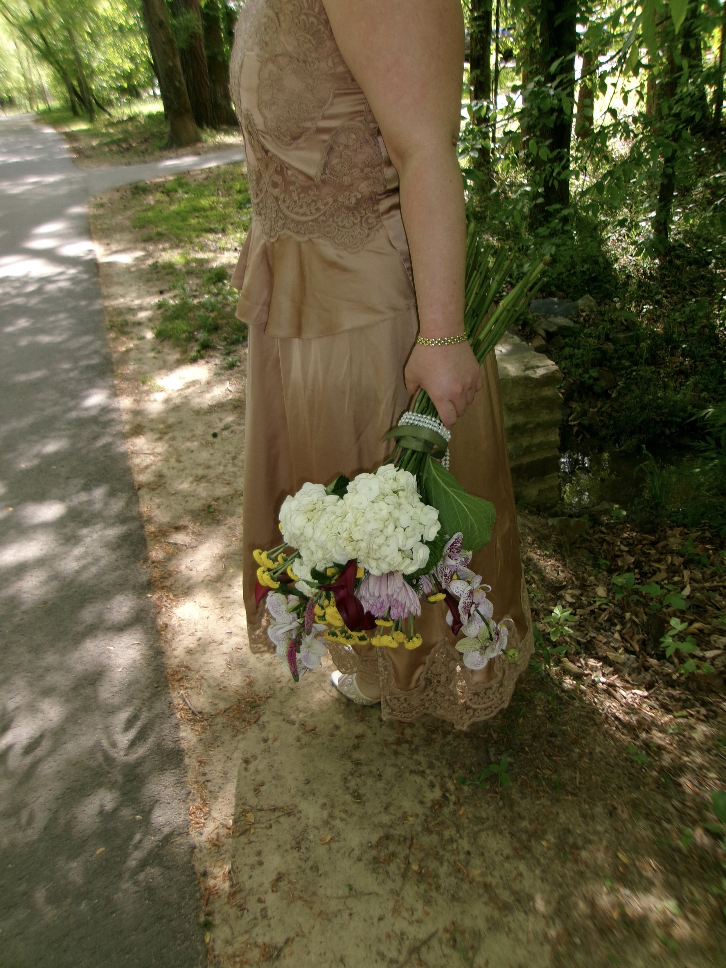 A woman holding a bouquet of white flowers and orchids, wearing a beige lace and satin dress, standing outdoors on a dirt path surrounded by trees.