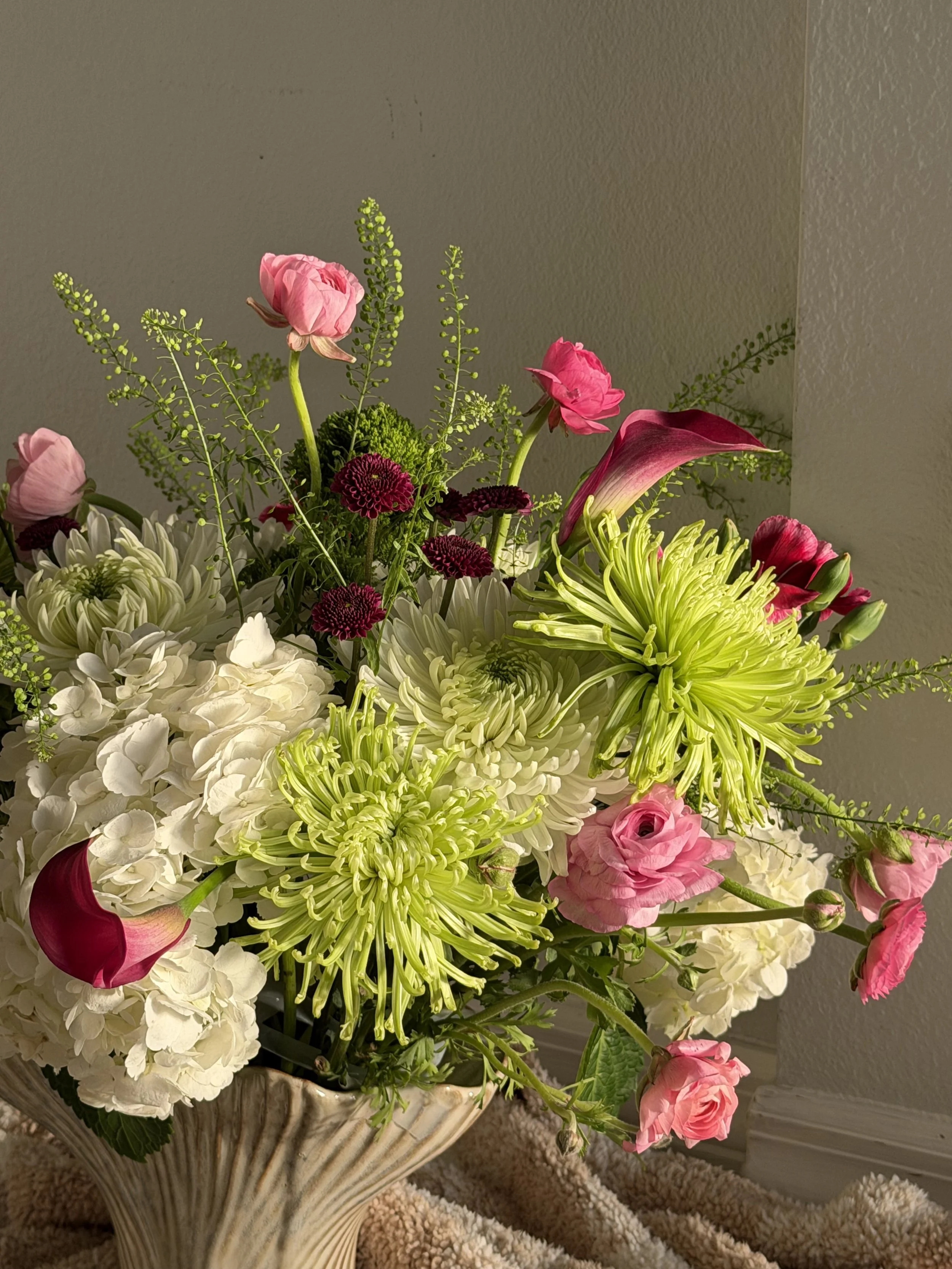 A floral arrangement in a decorative vase with pink, white, green, and burgundy flowers, including hydrangeas, dahlias, roses, and calla lilies, placed on a beige carpeted floor against a gray wall.