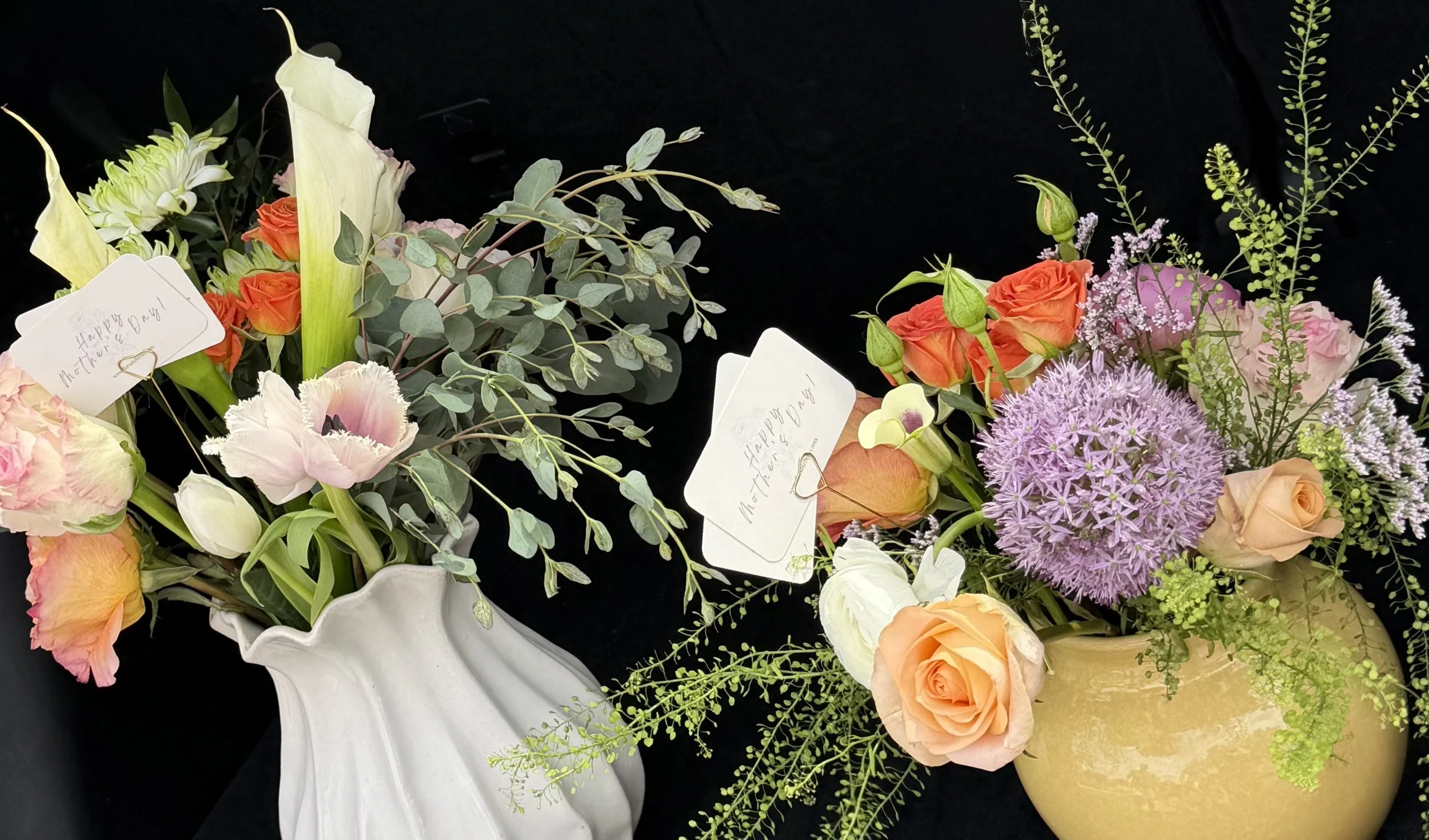 Arrangement of mixed fresh flowers including roses, lilies, lisianthus, and greenery in white and yellow vases, with small notes attached that read 'Happy Mother's Day!'