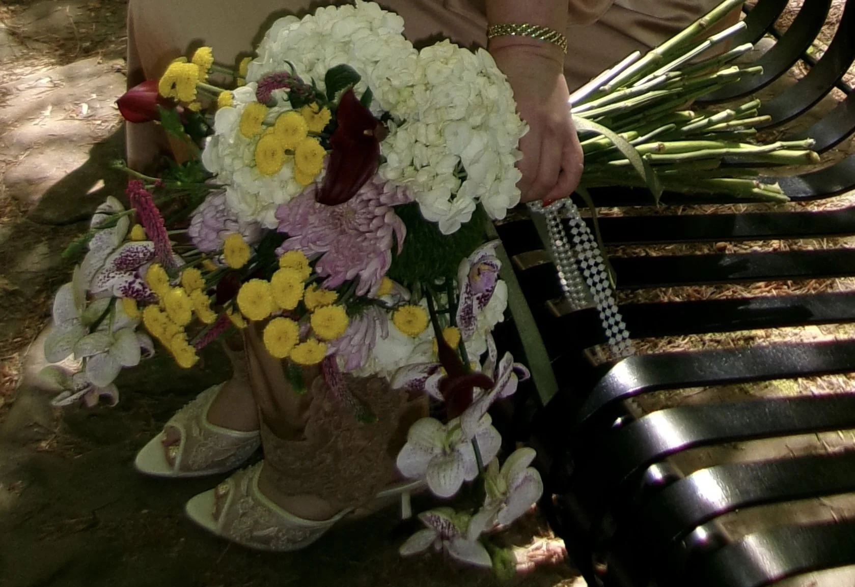 A person holding a bouquet of colorful flowers while sitting on a black park bench. The bouquet includes white, pink, yellow, and purple flowers, with some long green stems. The person is wearing a beige or light brown dress and white shoes, with one arm adorned with gold and pearl jewelry.