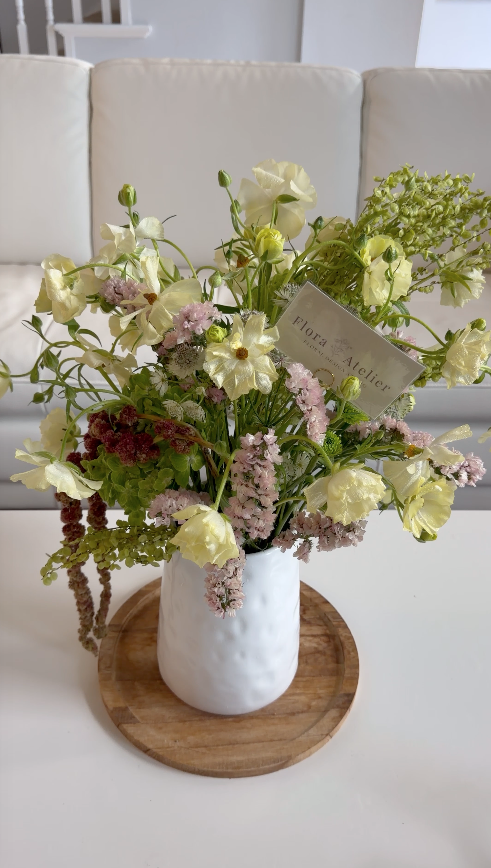 A white ceramic vase filled with a bouquet of cream, pink, and green flowers, placed on a round wooden tray on a white table, with a white wall and partial staircase in the background.