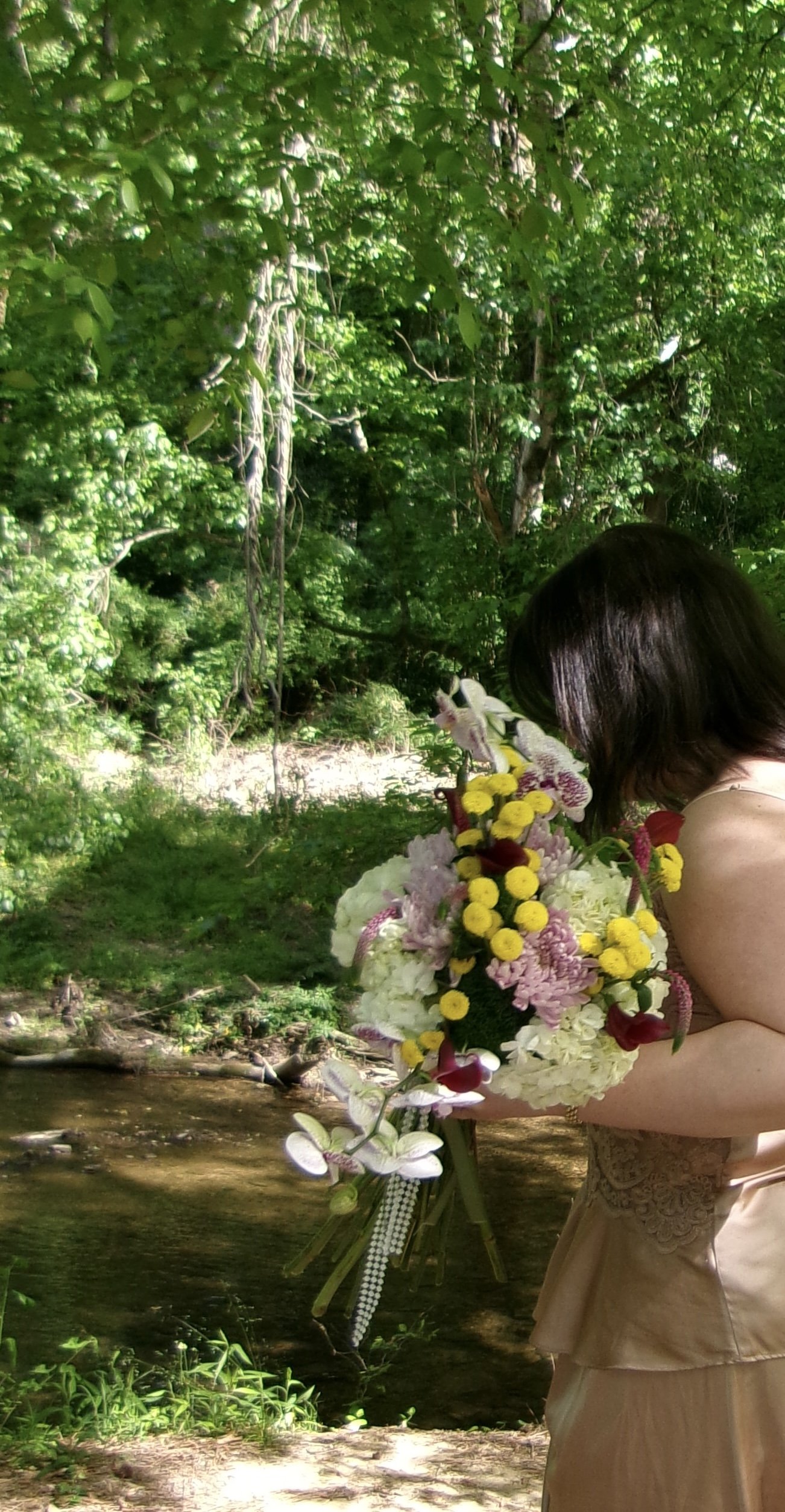 A woman in a beige dress is holding a large bouquet of colorful flowers near a stream in a lush green forest.