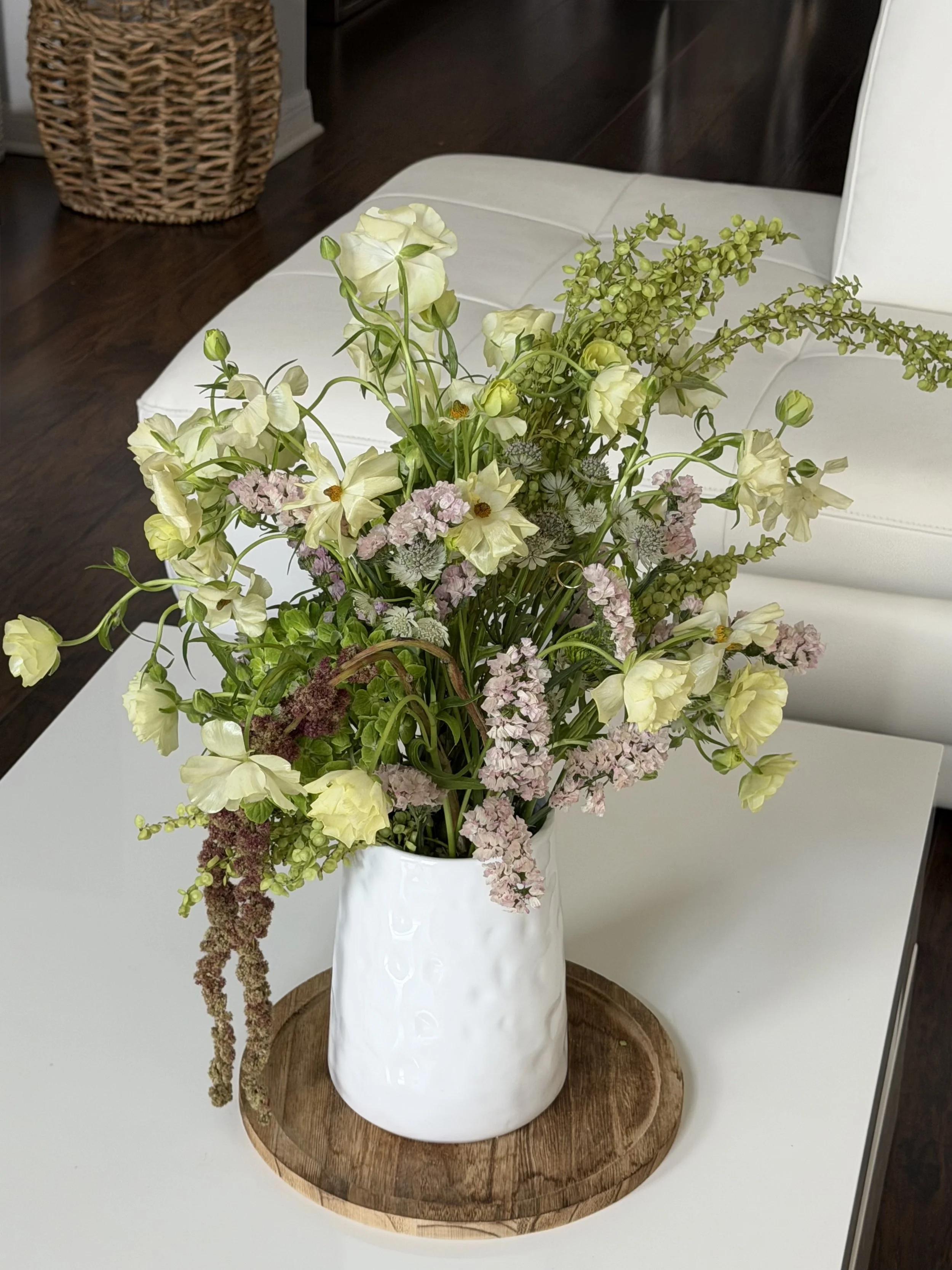 A white ceramic vase filled with a variety of pastel-colored flowers, including white roses, light purple, and green floral blooms, placed on a round wooden tray on a white surface.