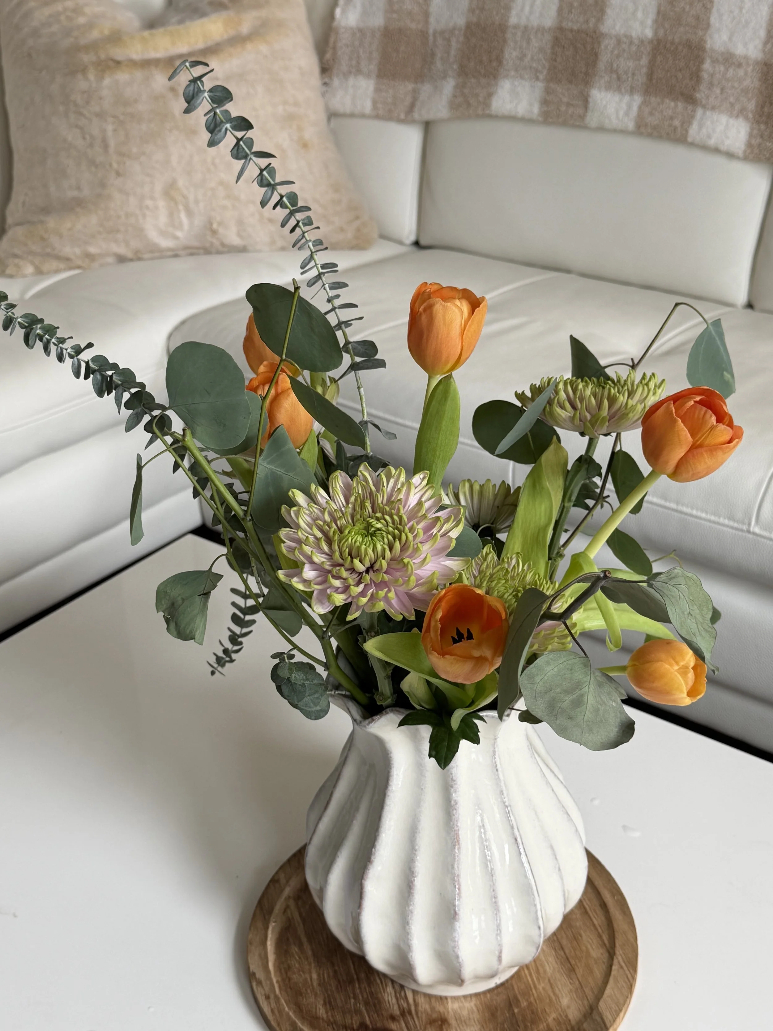A white ceramic scalloped vase holding a floral arrangement with orange tulips, green leaves, and greenery, placed on a round wooden tray on a white table in front of a white sofa with a beige pillow and a pink and beige plaid blanket.