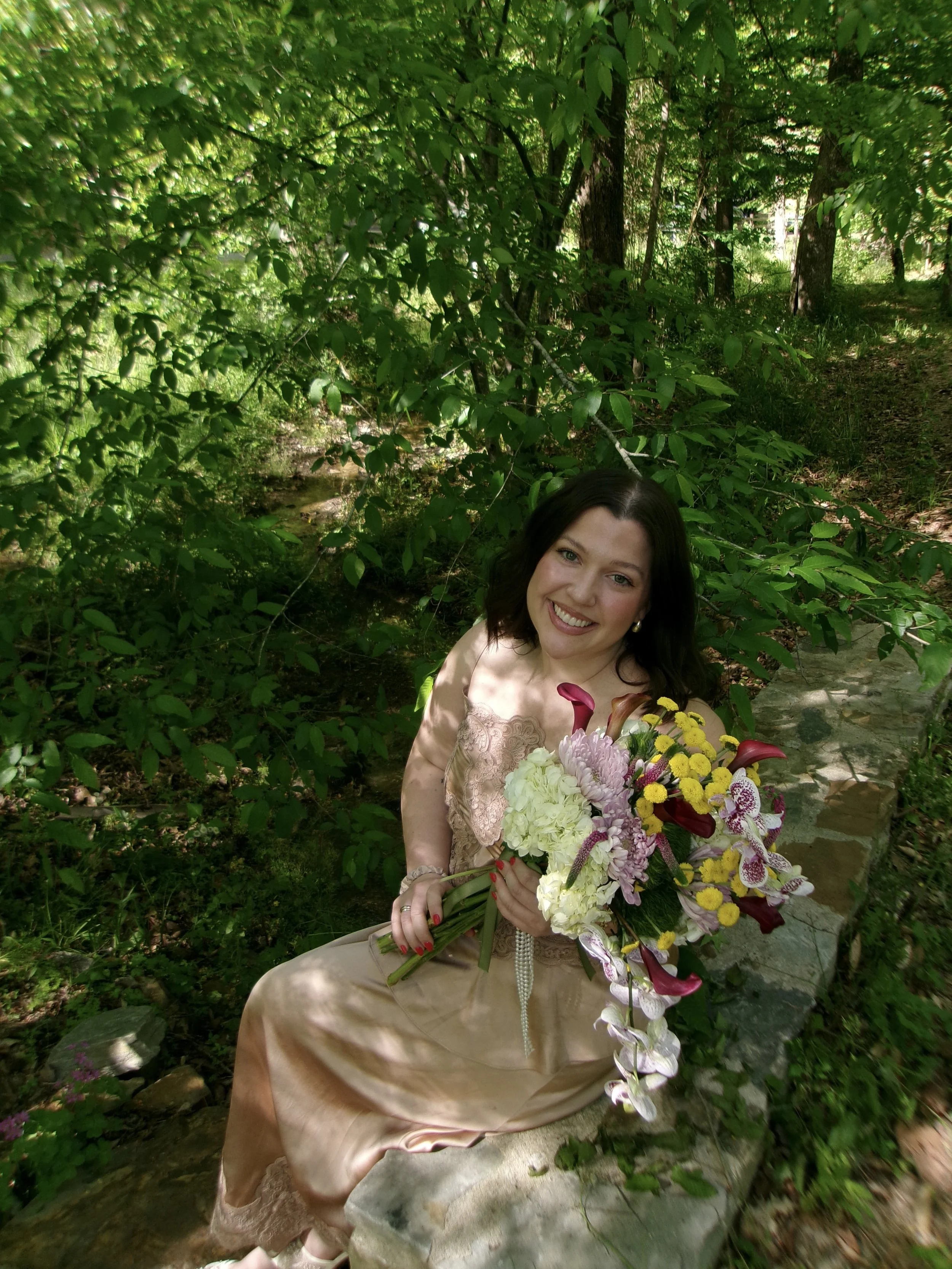 A woman with dark hair and fair skin smiling while holding a large bouquet of colorful flowers, sitting outdoors in a lush green forest.