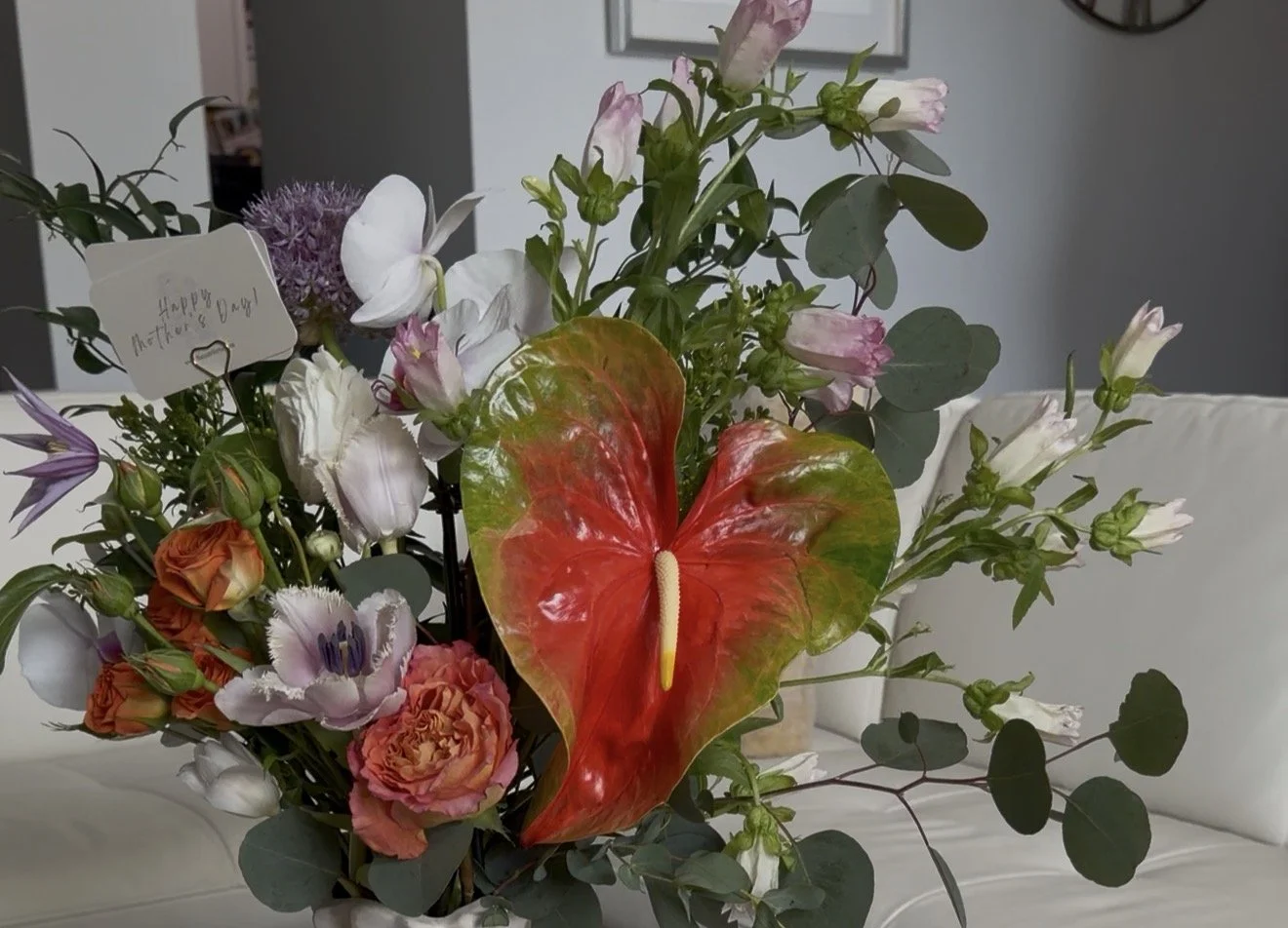 A floral arrangement in a vase with various colorful flowers, including a red and green anthurium, delicate white and purple blooms, orange roses, and greenery. There is a small white card with a handwritten message attached to the arrangement, reading 'Happy Mother's Day!'