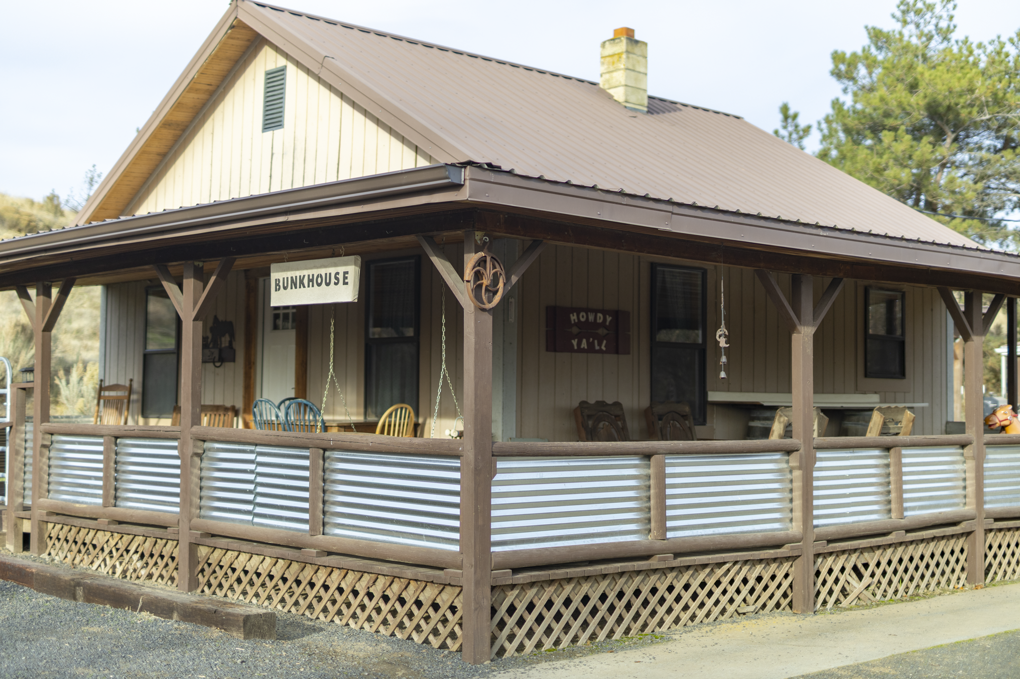 A wooden house with a porch that has colorful chairs and hanging decorations. There are signs reading 'BUNKHOUSE' and 'HOWDY YA'LL' on the house. The house has a metal roof and is surrounded by trees.