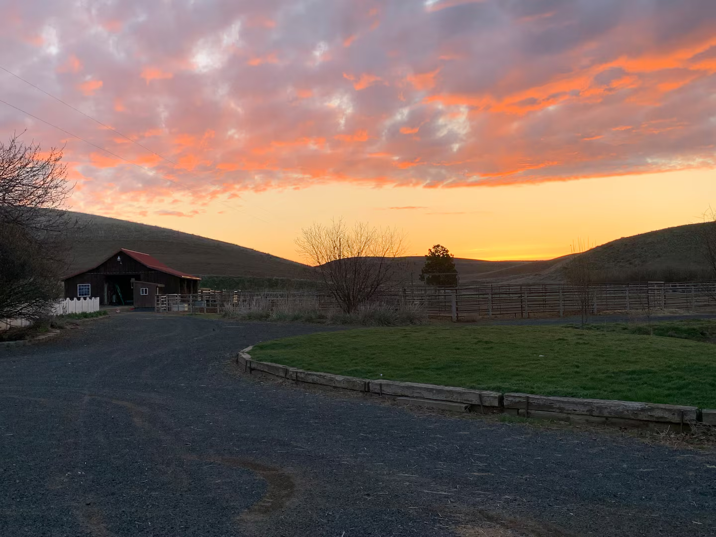 Sunset over a rural farm landscape with a barn, trees, grassy area, and rolling hills, under a colorful sky with orange and purple clouds.