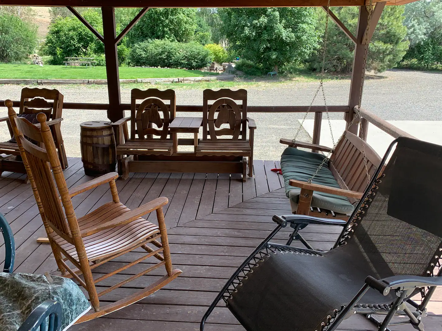 Covered porch with wooden furniture including a rocking chair, a set of chairs with a small table, a wooden bench with backrests, a hanging swing with cushions, and a lounge chair. The porch overlooks a driveway and a lush green yard with trees and bushes.