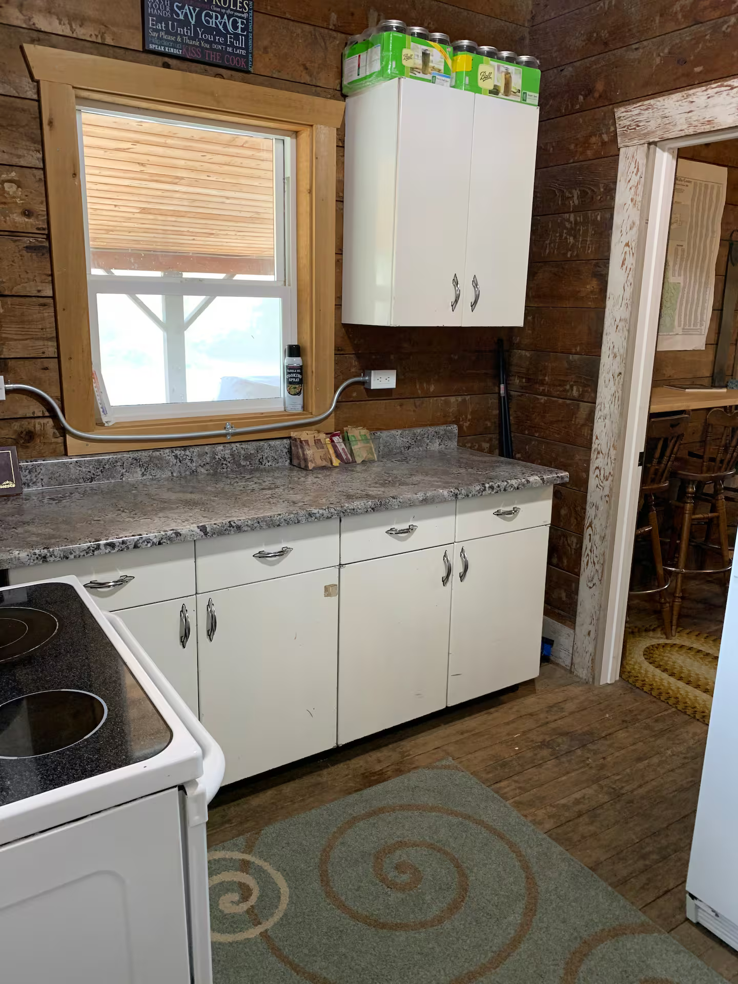 A rustic kitchen with wooden walls, a window above the sink, white cabinets, a grey speckled countertop, and a stove. There are several cans of cooking spray on top of a tall white cabinet, and a colorful woven rug on the wooden floor.