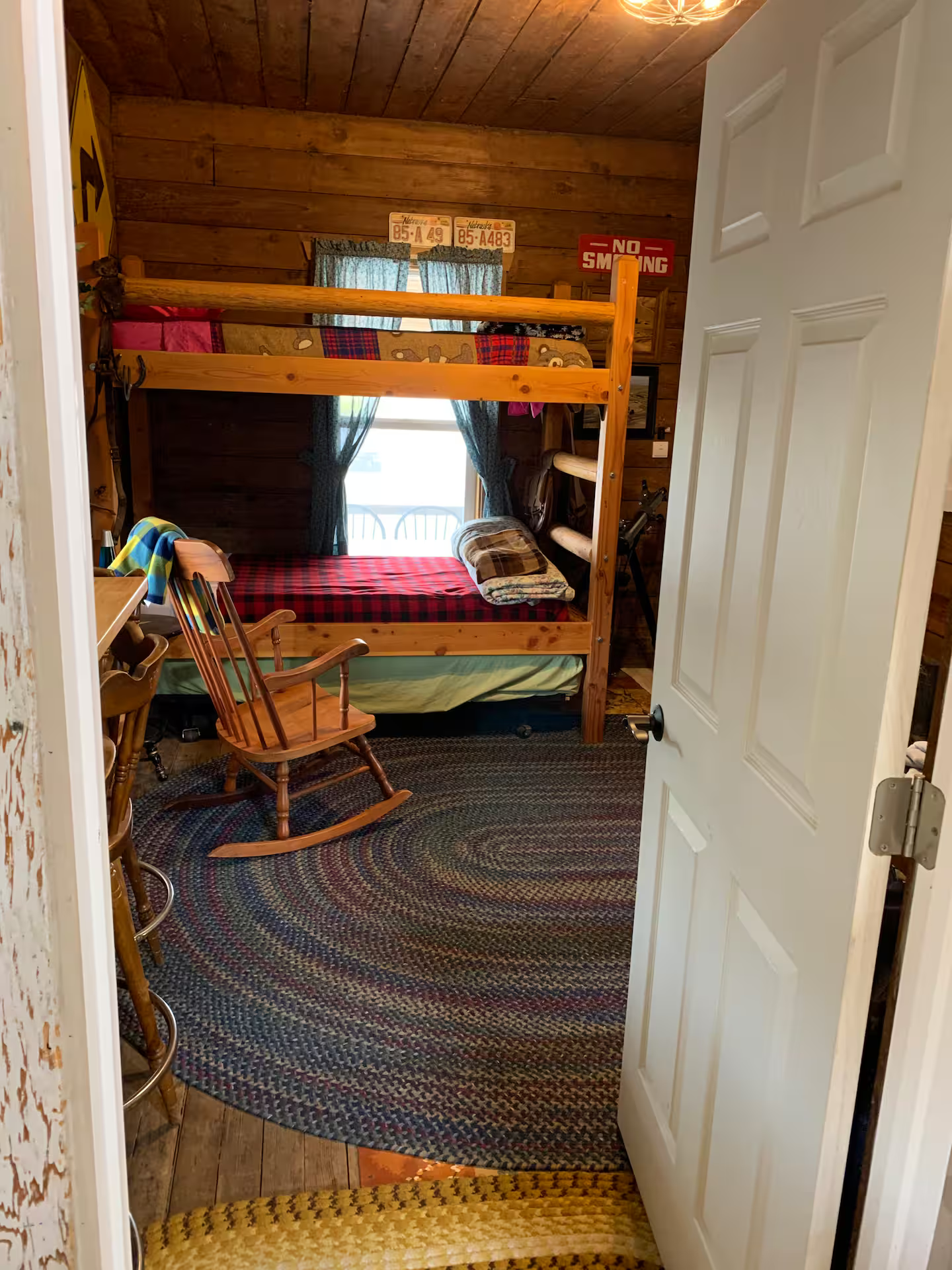 A cozy bunkhou bedroom with a bunk bed, a wooden rocking chair, and various decorations on the walls, including license plates and signs. The room features wooden walls and ceiling, a window with curtains, and a multicolored braided rug on the floor.