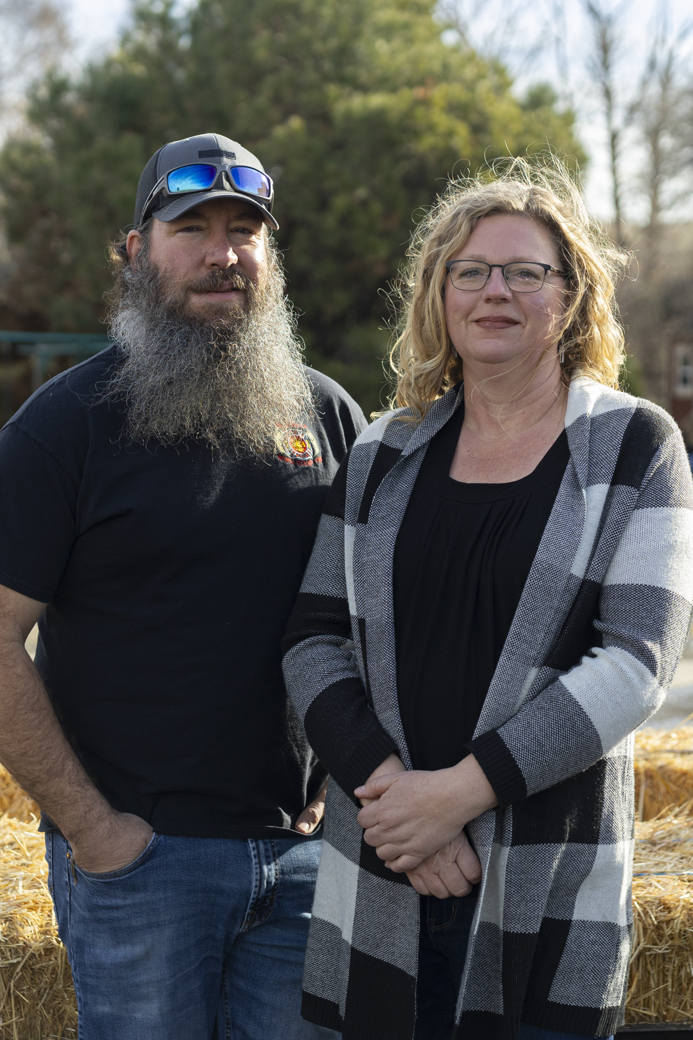 A man with a long gray beard wearing sunglasses and a black t-shirt, standing next to a woman with curly blonde hair and glasses, wearing a black top and a gray plaid cardigan, outdoors near hay bales.