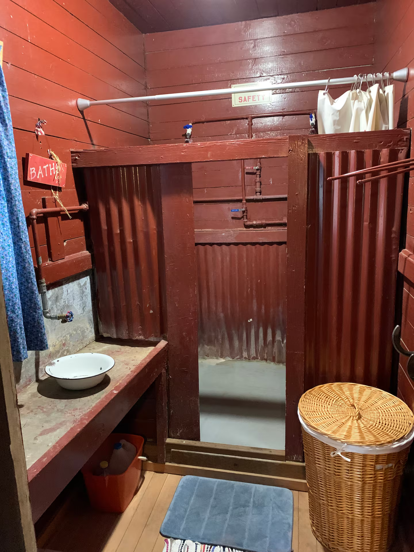 Small indoor bathroom with wooden walls, a shower area with a corrugated metal wall, a wooden countertop with a white bowl, a trash can, a blue bath mat, and a woven laundry basket.