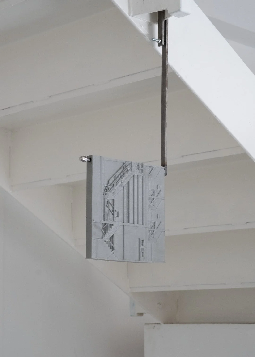 A metal sculpture of an industrial staircase and piping hangs from the ceiling, secured by a metal rod with bolts in a white-walled room.
