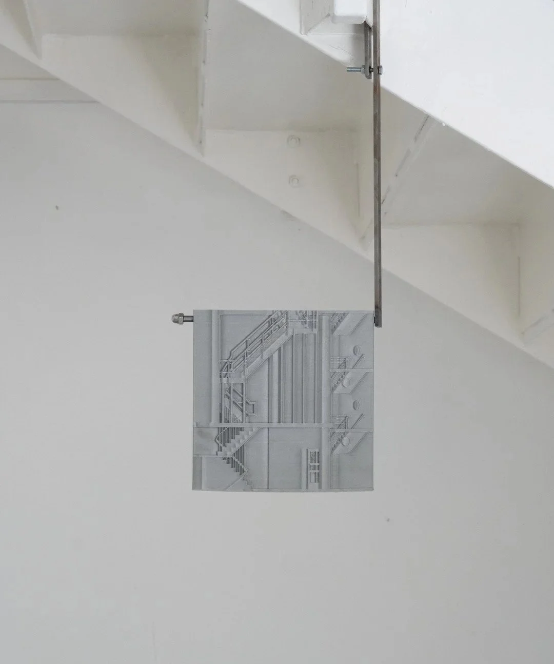 A metal sculpture depicting a detailed industrial staircase and building structure, mounted on a white wall under a staircase.