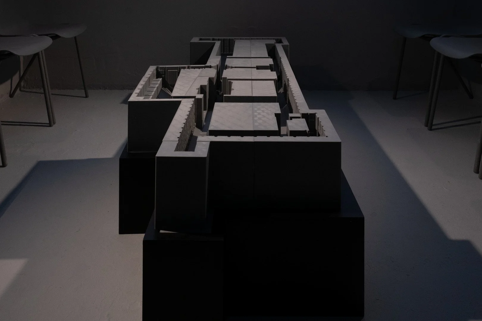 A large architectural model or structure on a black pedestal, viewed from the front, in a dimly lit room with chairs on either side.
