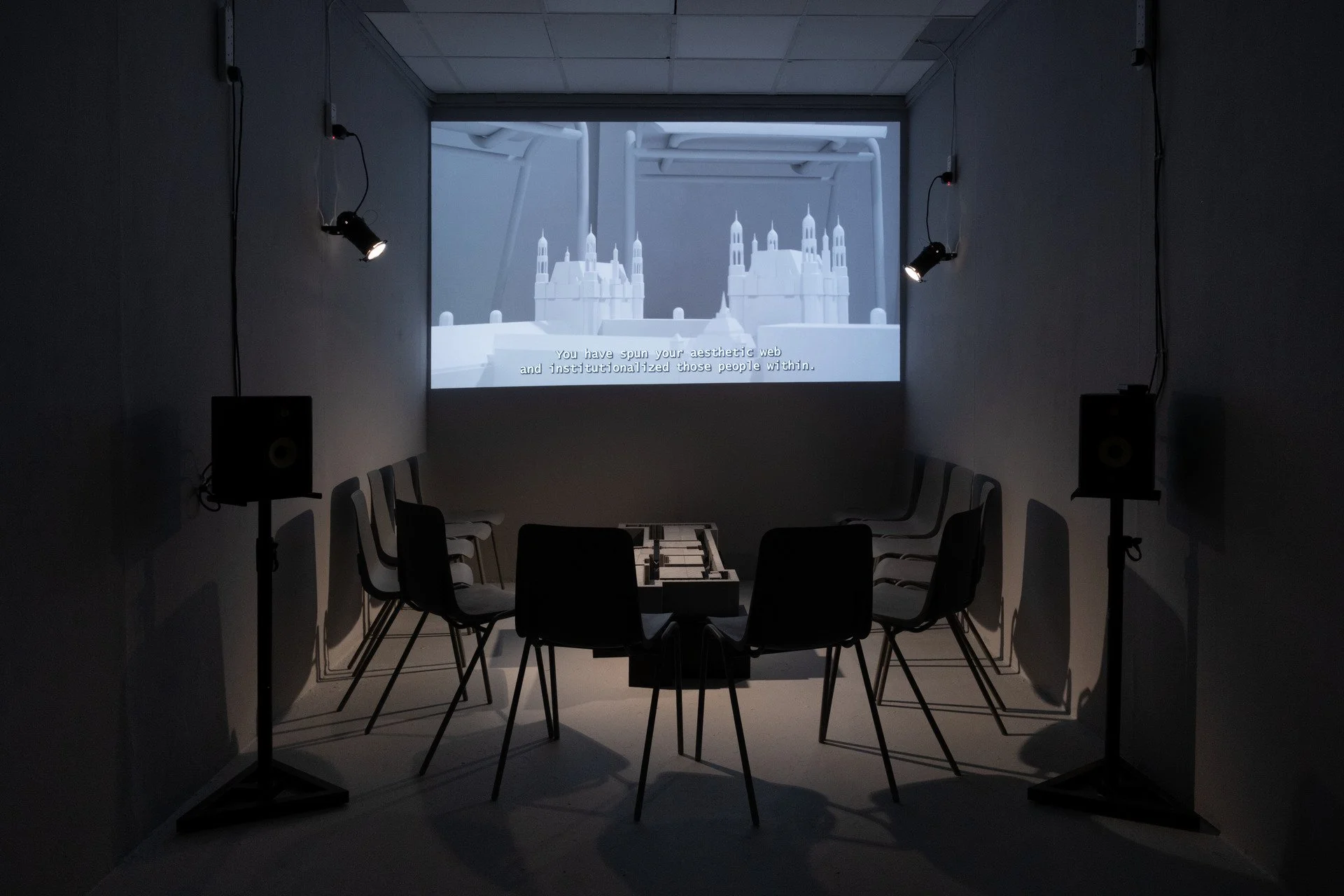 An art gallery or exhibition room with chairs arranged in a circle around a small table, facing a large screen displaying a digital image of a white castle with tall spires. The room has dark walls, chairs, and two spotlights on stands illuminating t