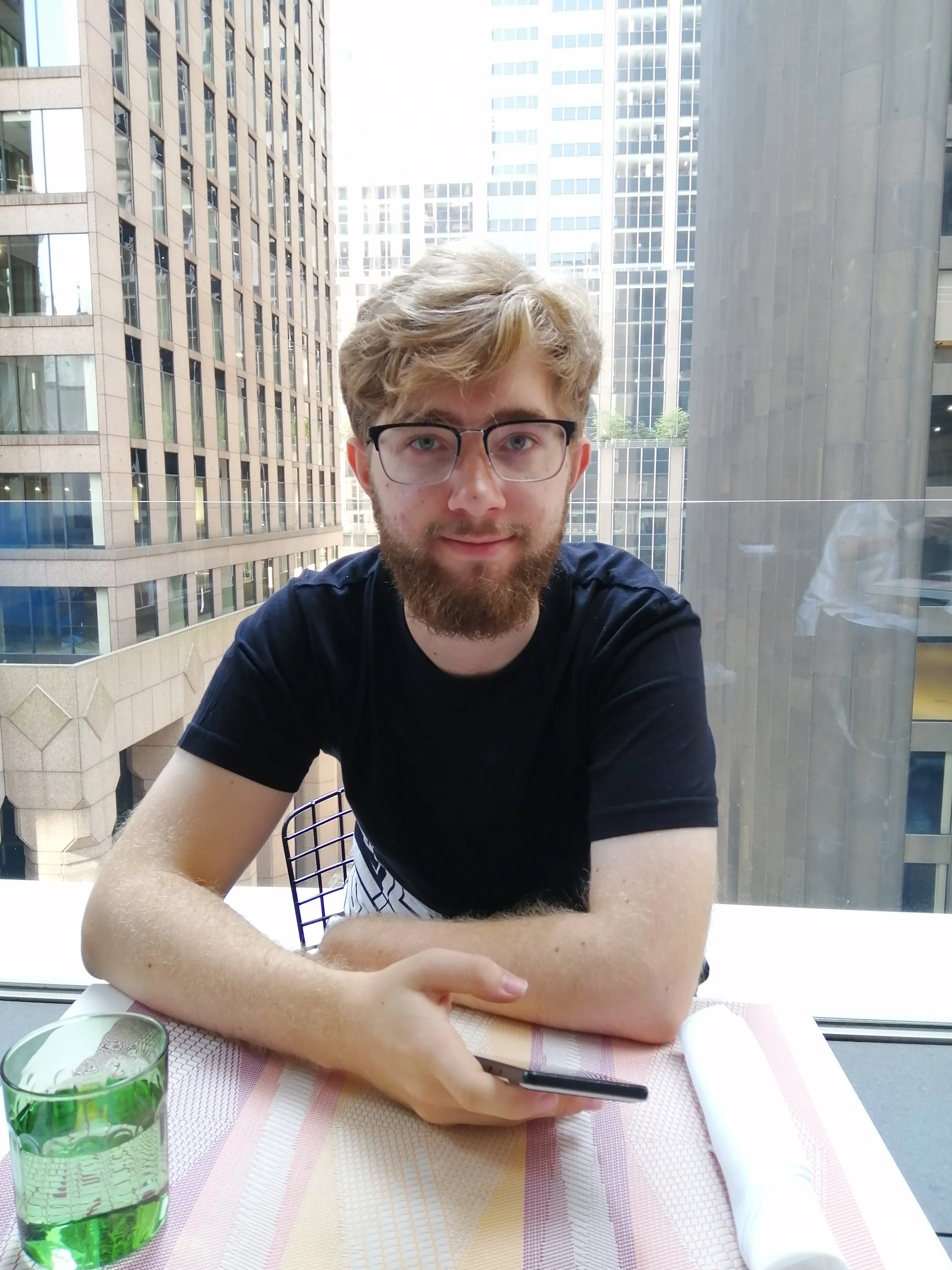 A young man with light brown curly hair, a beard, and glasses, is sitting at a table on a balcony of a high-rise building with a cityscape of tall commercial buildings in the background. He is wearing a dark T-shirt and has his arms crossed on the table. In front of him are a green glass of water, a pen, a napkin, and a white rolled-up paper or napkin.