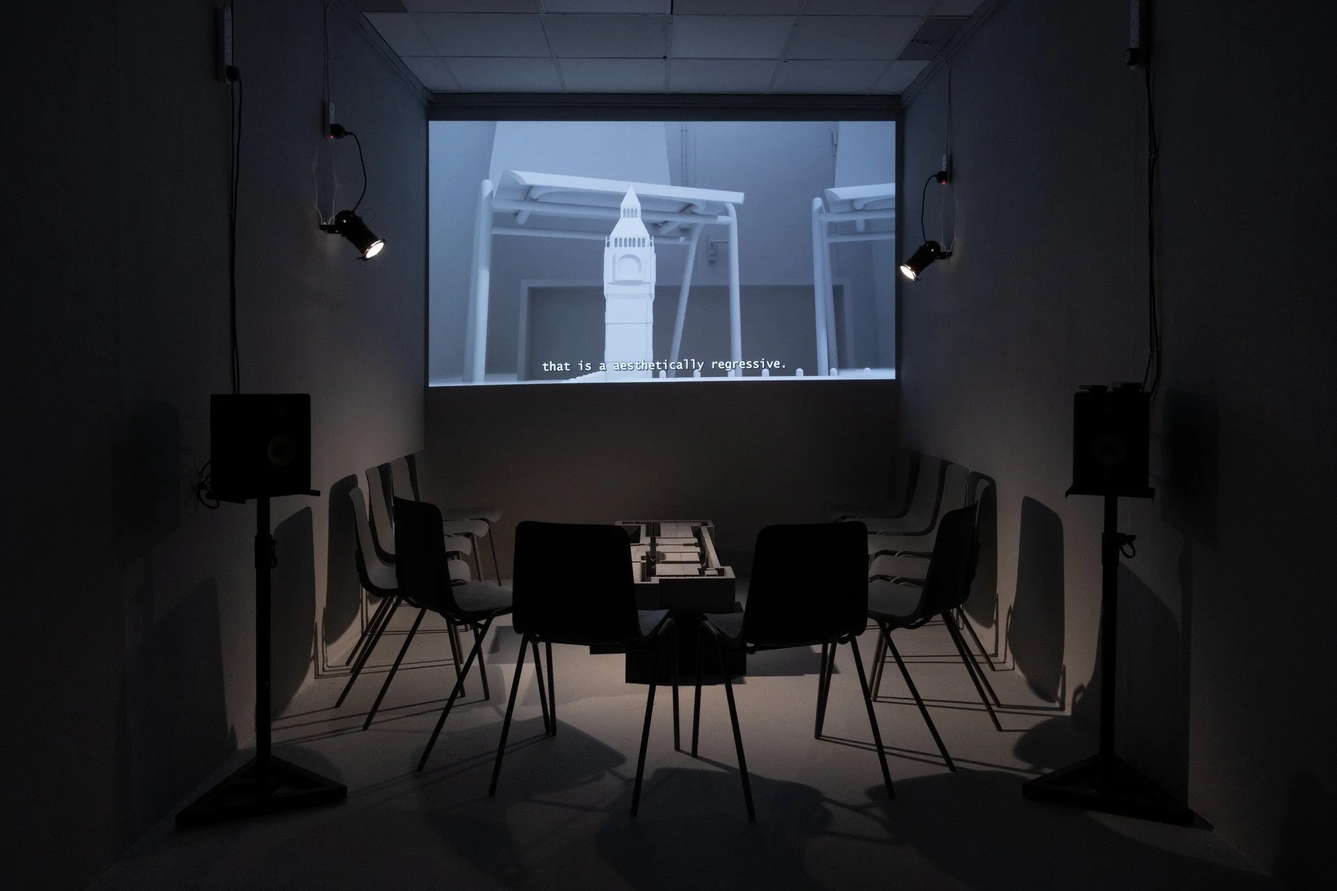 Dark room with chairs arranged in a circle, speakers on stands, and a projection screen displaying a video of digitally remodelled Westminster Palace.