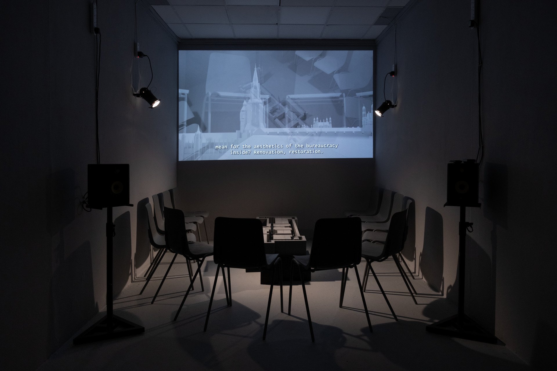 A dimly lit room with chairs arranged in a circle around a rectangular table, speakers on stands on either side, and a projection screen showing architectural design with subtitles reading "mean for the aesthetics of the bureaucracy inside? Renovatio