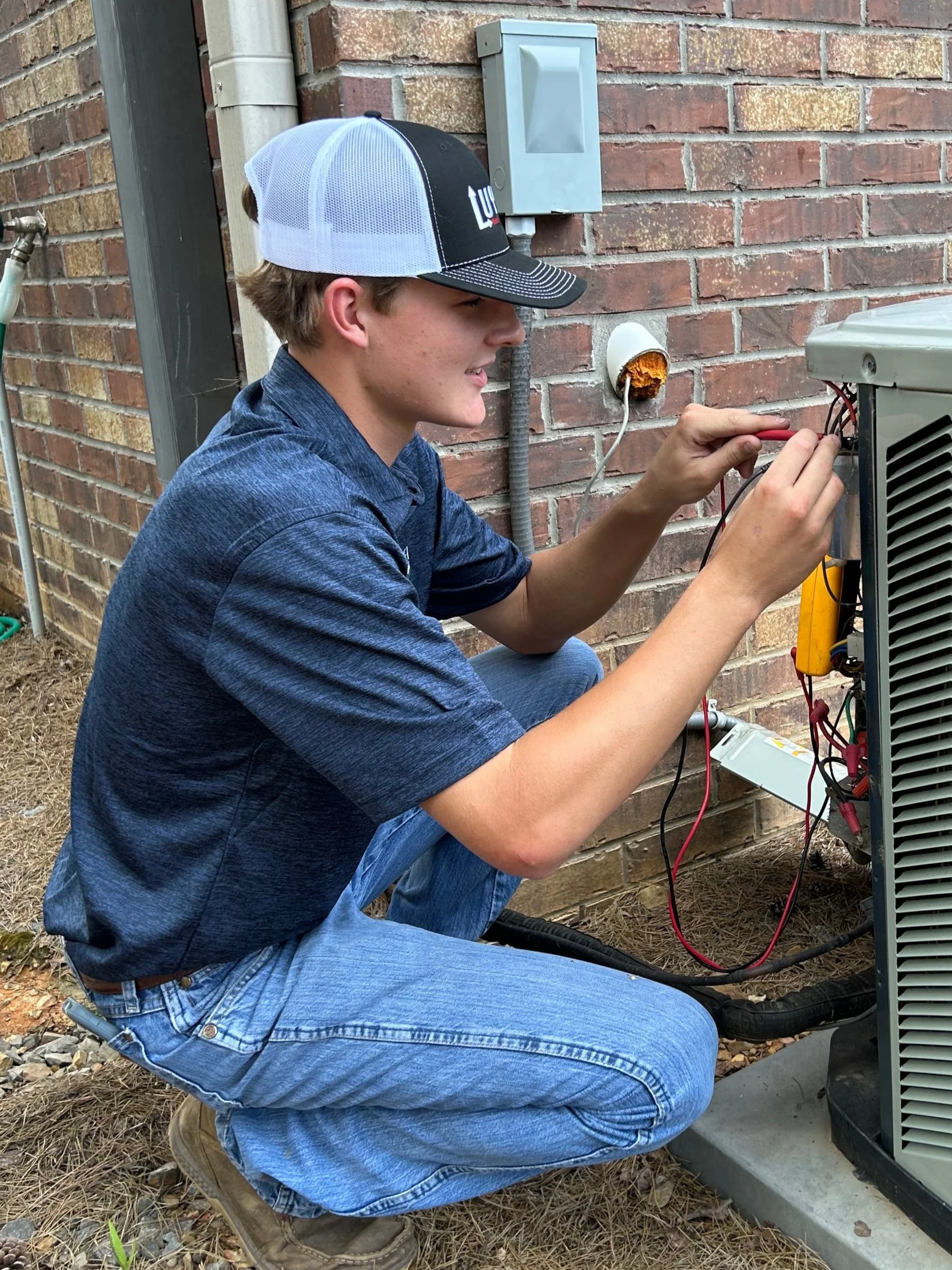 A young man wearing a blue shirt, jeans, and a trucker hat is working on an outdoor HVAC unit using a multimeter for testing wiring connections.