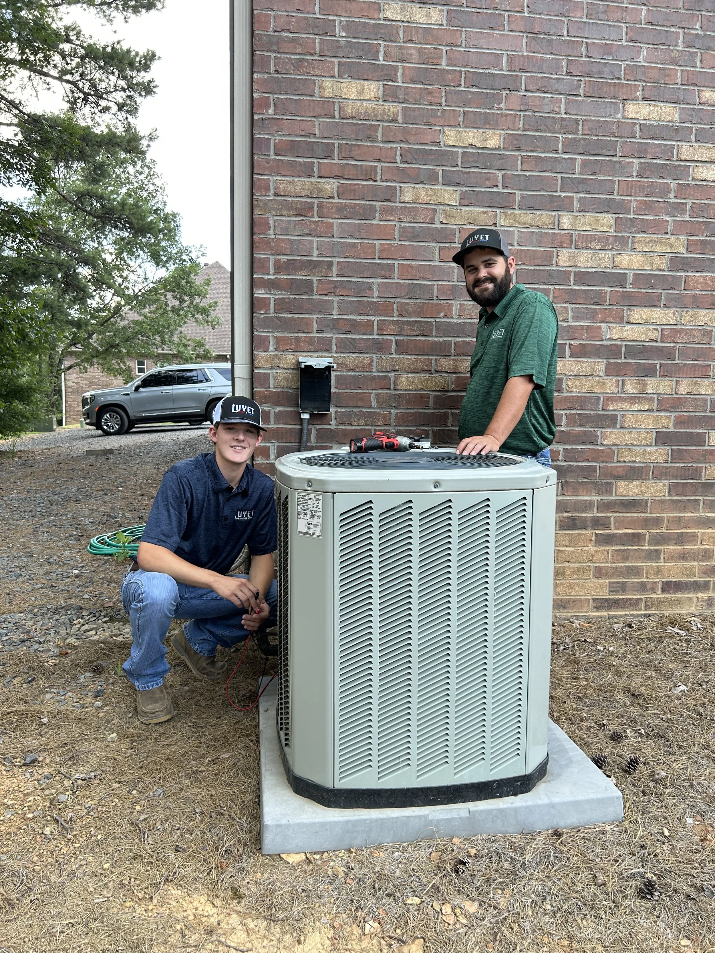 Two men working on an outdoor air conditioning unit next to a brick wall. Both are wearing caps and work shirts with the logo 'LUYET'. One man is standing, the other is crouching with tools and wires.