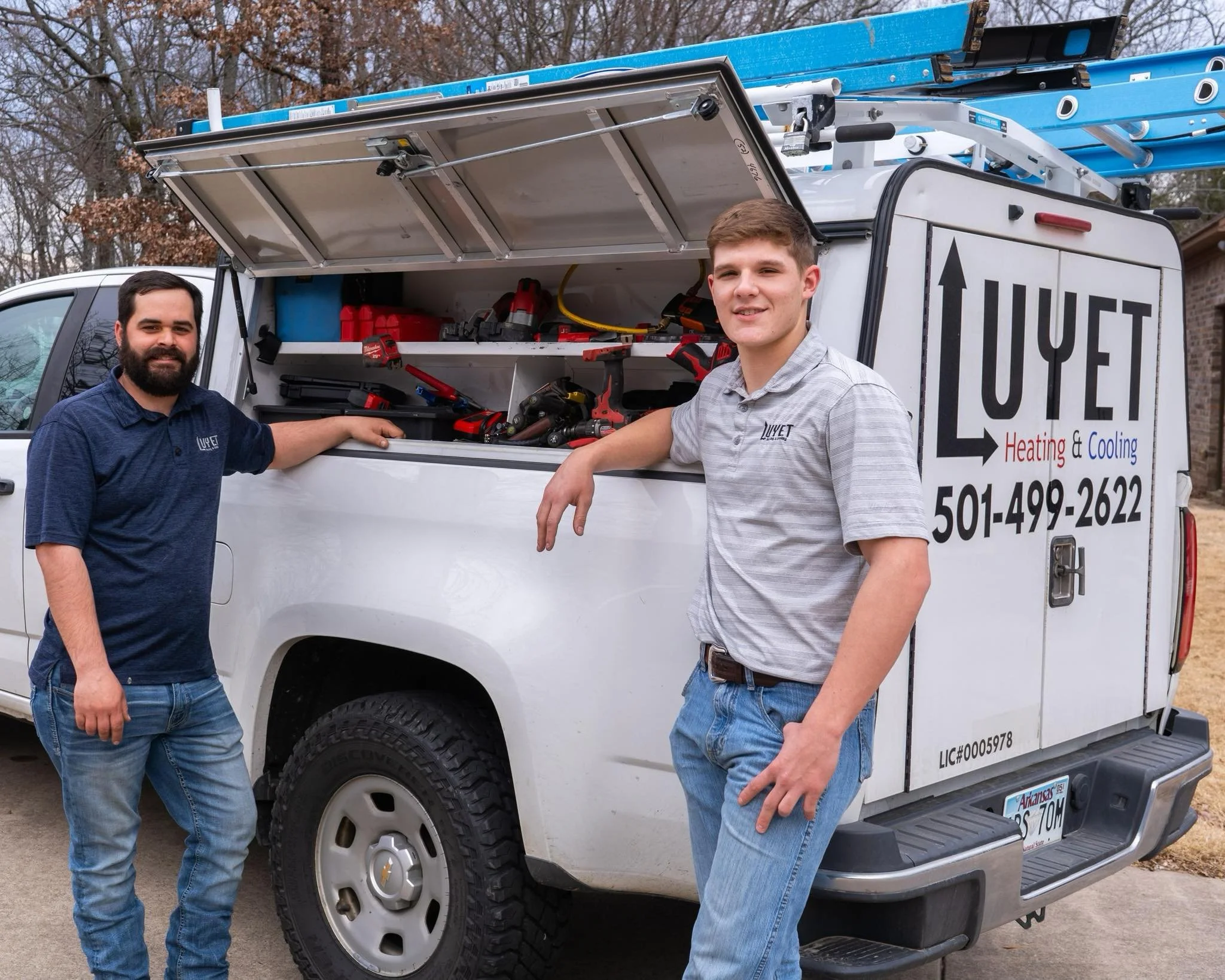 Two men standing beside a white service truck with the text 'LUYET Heating & Cooling' and a phone number on the side. The truck's open compartment contains tools.