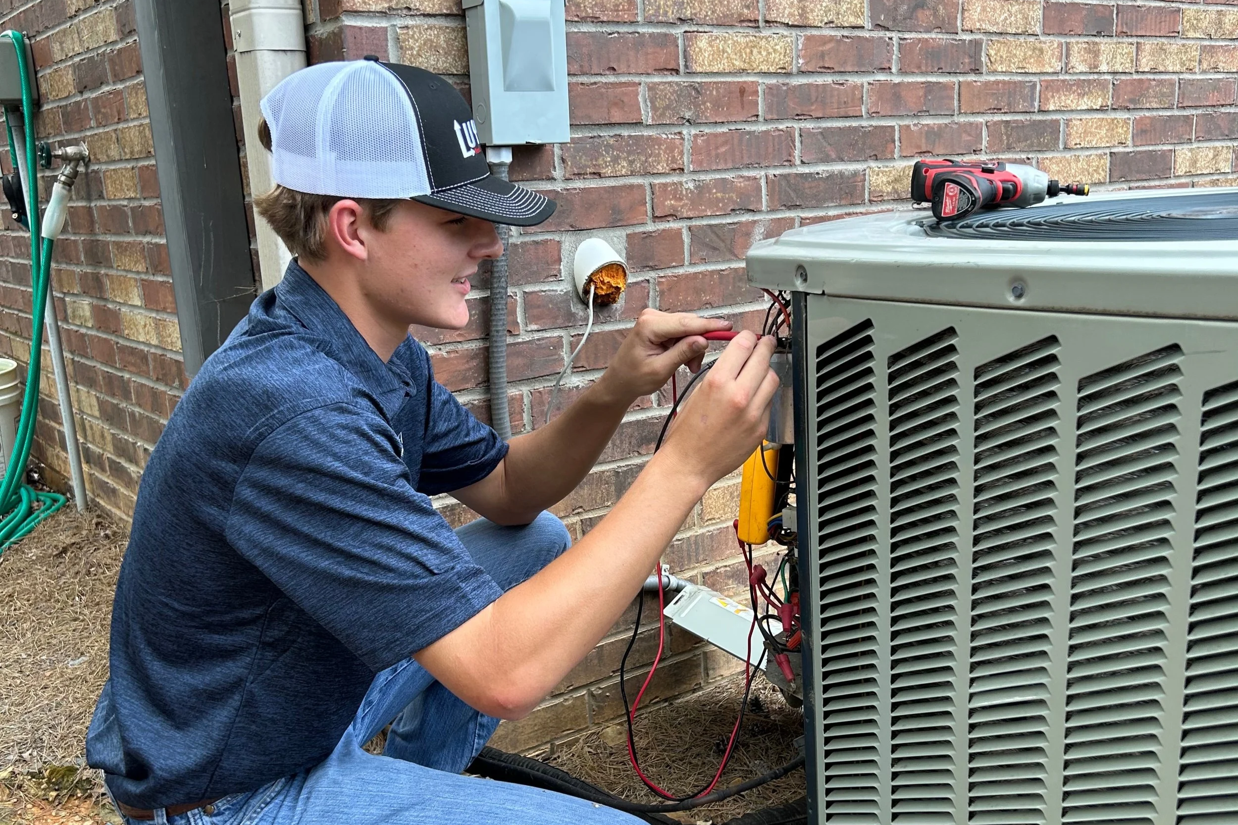 A young man wearing a blue shirt and a baseball cap is working on an air conditioning unit outside a building. He is using tools to repair or service the unit, with various wires and equipment around him.