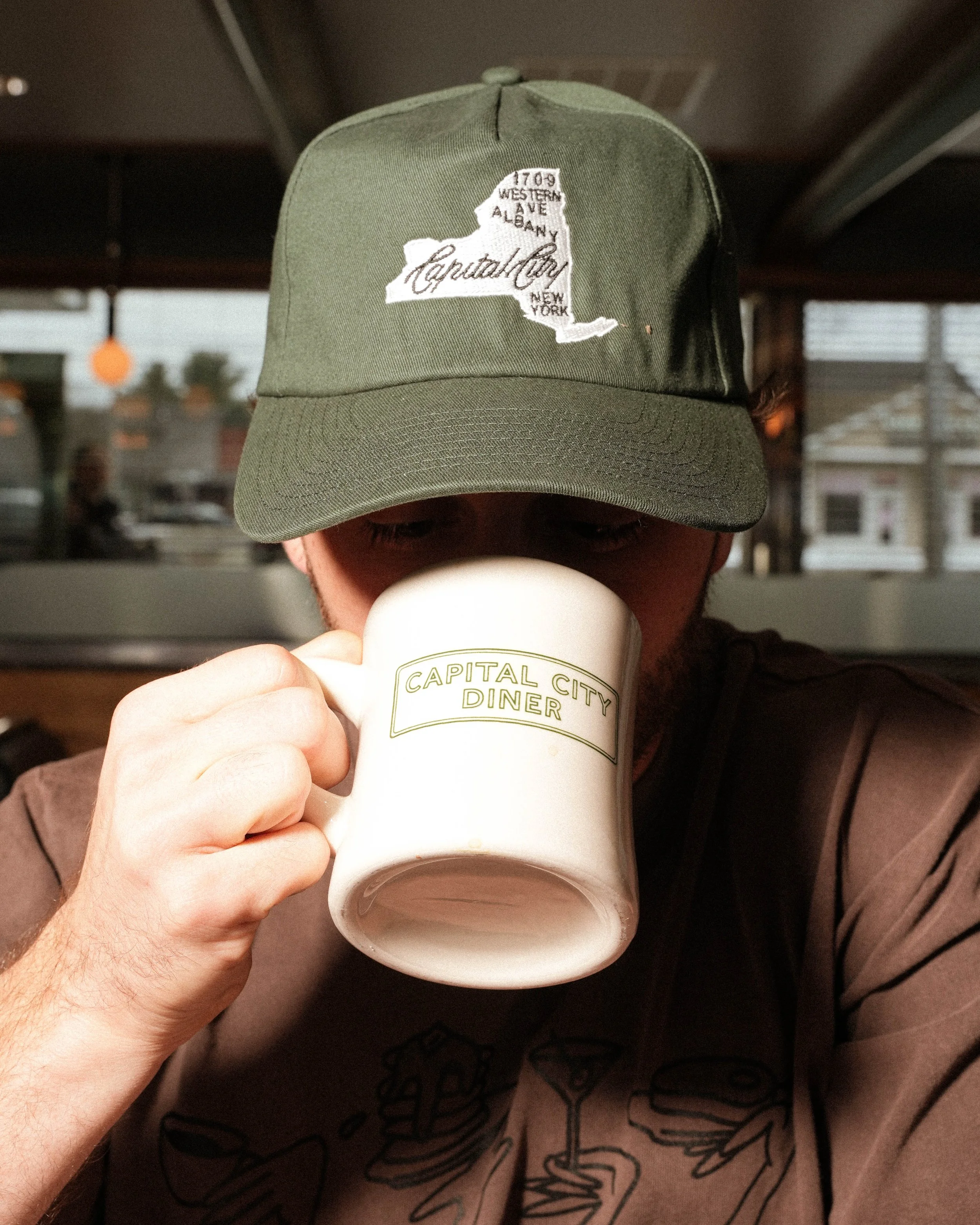 Person holding a white cup labeled 'Capital City Diner' while wearing a green baseball cap with an embroidered map of New York state and text indicating Albany, Western Ave, and Capitol City.