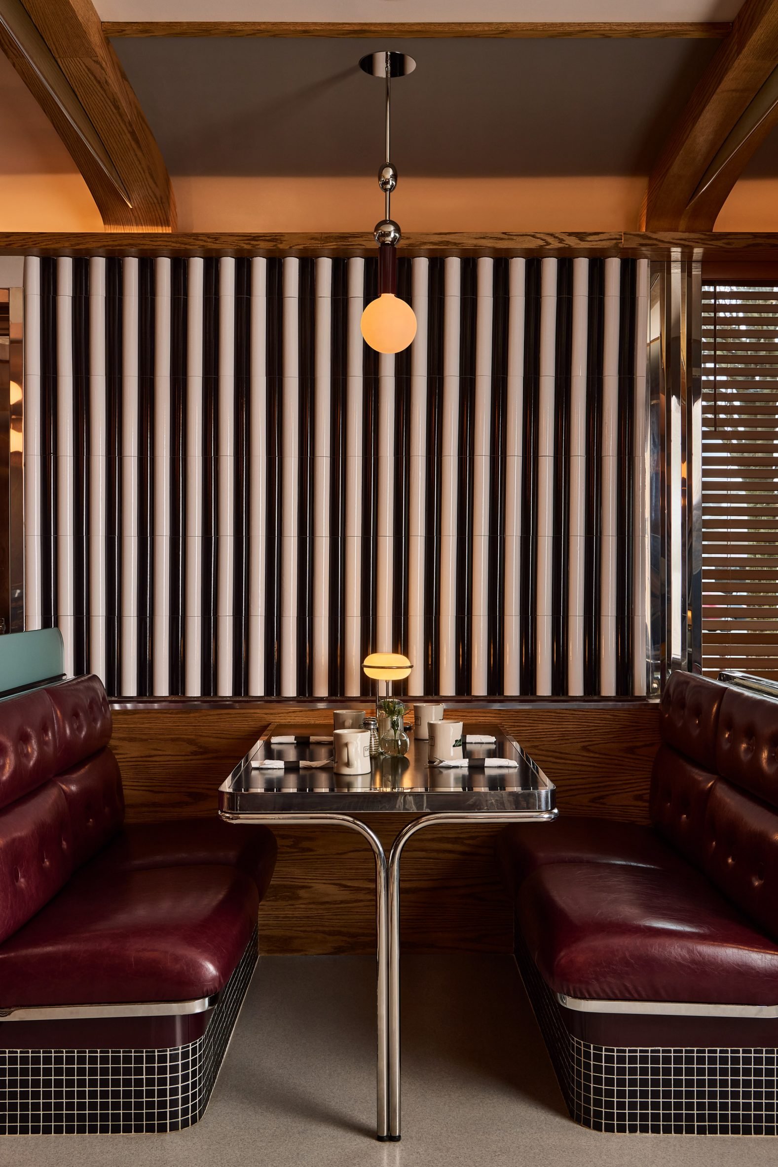 Interior of a retro-style diner booth with red leather seating, a glass-topped table, and a modern pendant light hanging above. Wooden accents and louvered window slats are visible in the background.