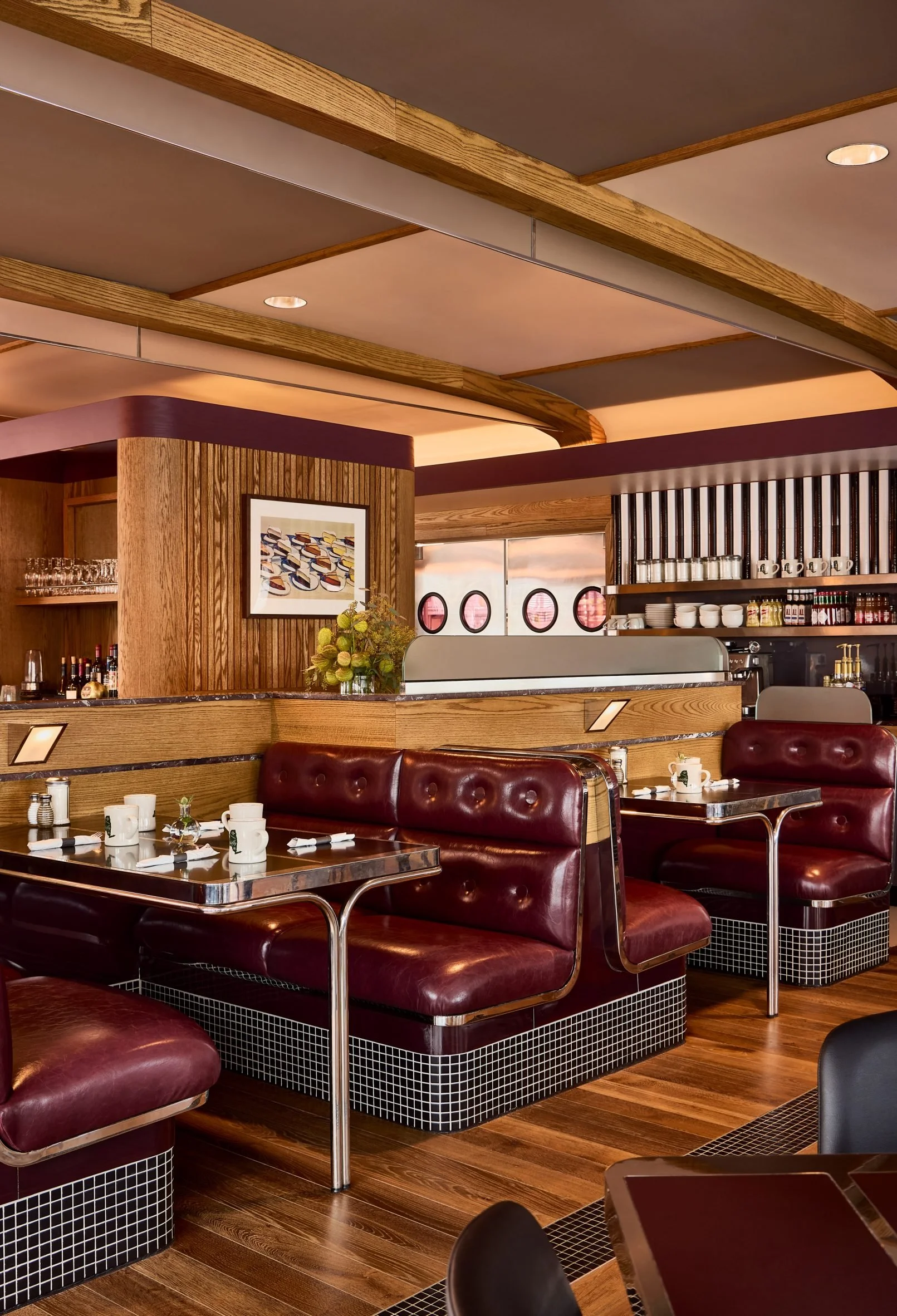 Inside a retro-style diner with burgundy leather booths, wooden accents, and a bar area with shelves of cups and glasses. Tables are set with cups, utensils, and small vases. Decorative artwork and patterned tile bases are visible.