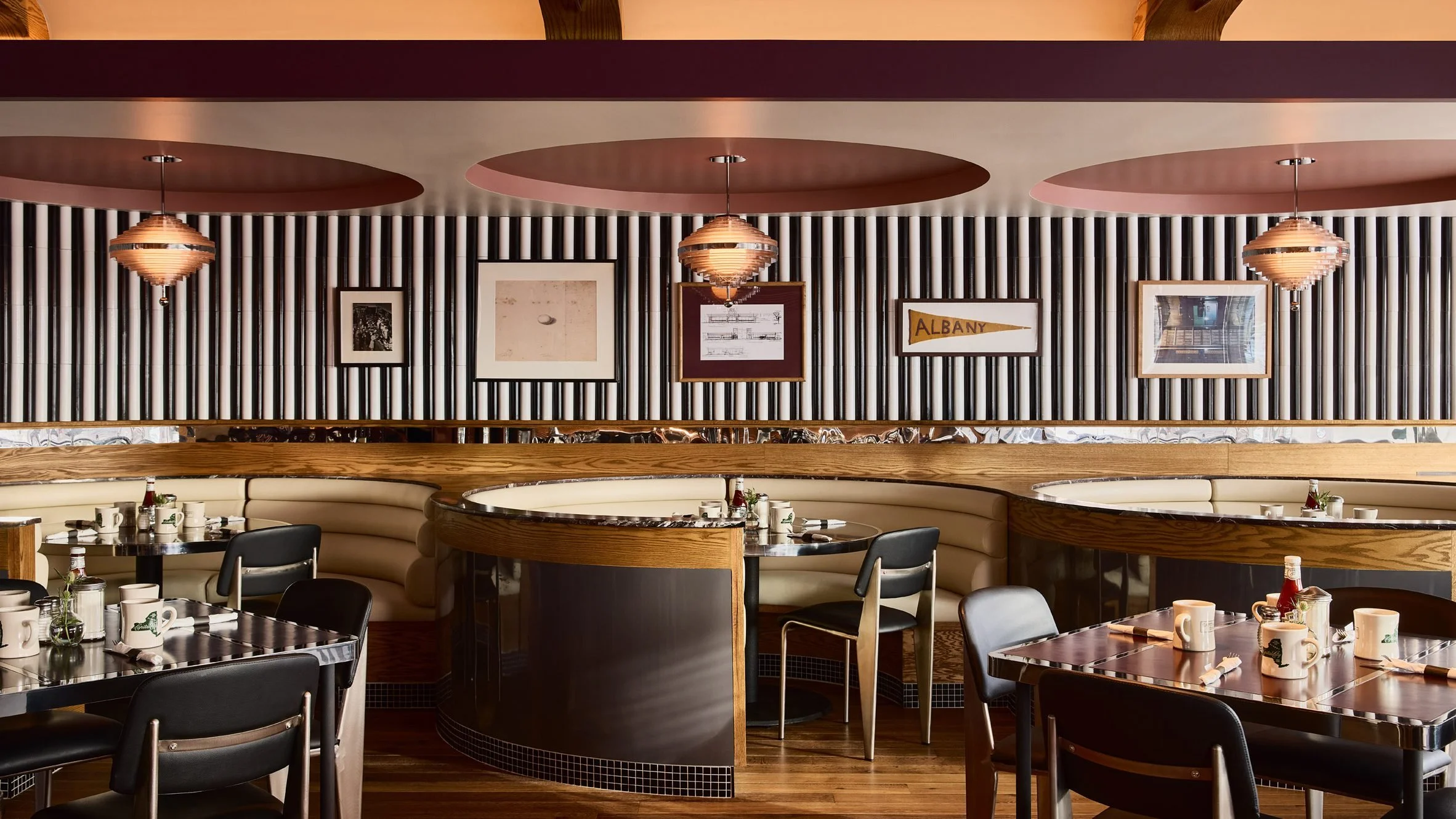Interior of a restaurant with black tables, white coffee mugs, and small condiment bottles, beige curved booths, wooden accents, framed pictures on a wall with black and white stripes, hanging pendant lights, and a sign that says "Albany".