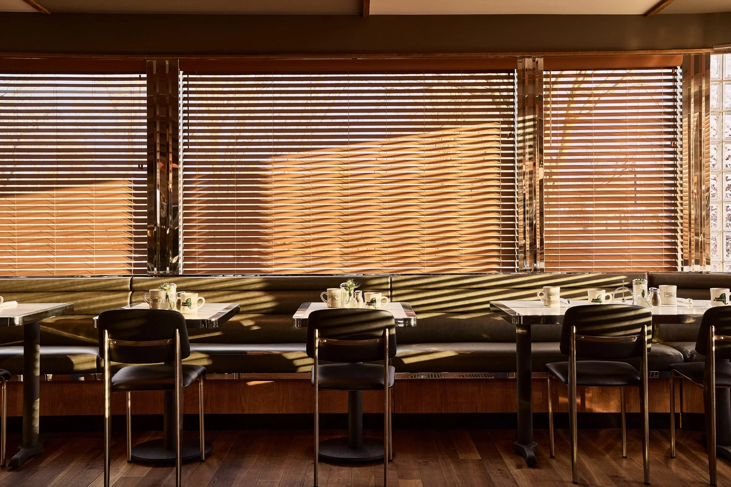 Interior of a restaurant or cafe with wooden blinds covering large windows, tables set with cups and utensils, and sunlight creating shadows on the tables.