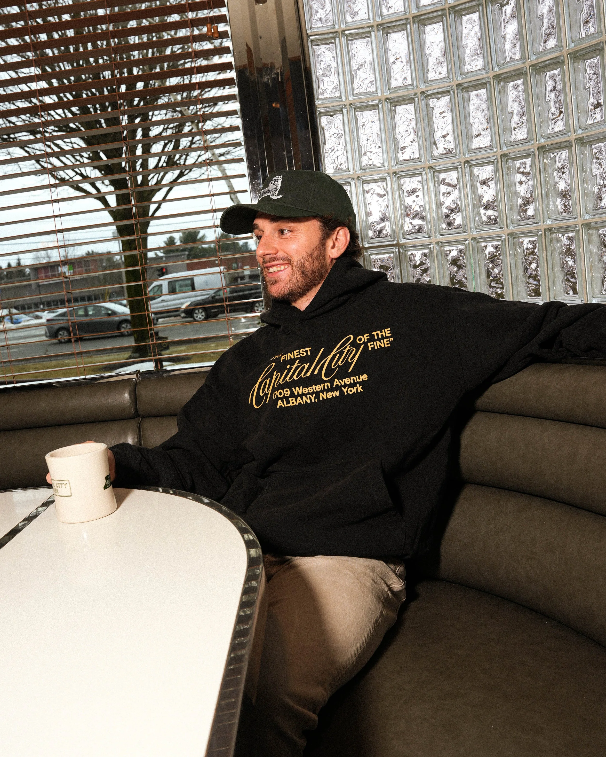 A man is sitting in a diner, smiling, holding a white mug. He wears a black cap and a black hoodie with gold text. The background shows large glass blocks and window blinds, with a view of parked cars outside.