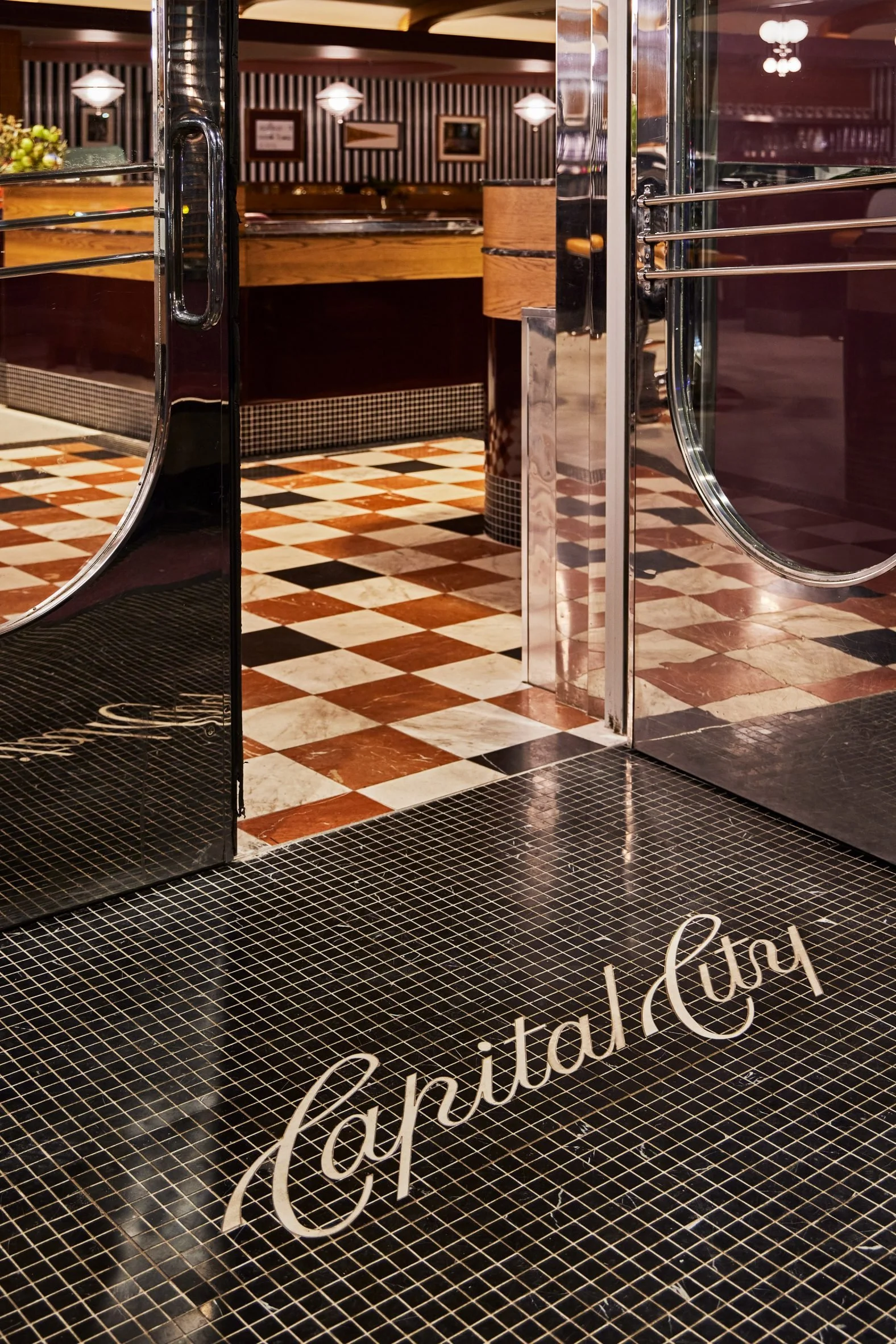 Open glass door showing the interior of a restaurant with a checkered black and white tile floor and elegant lighting fixtures.