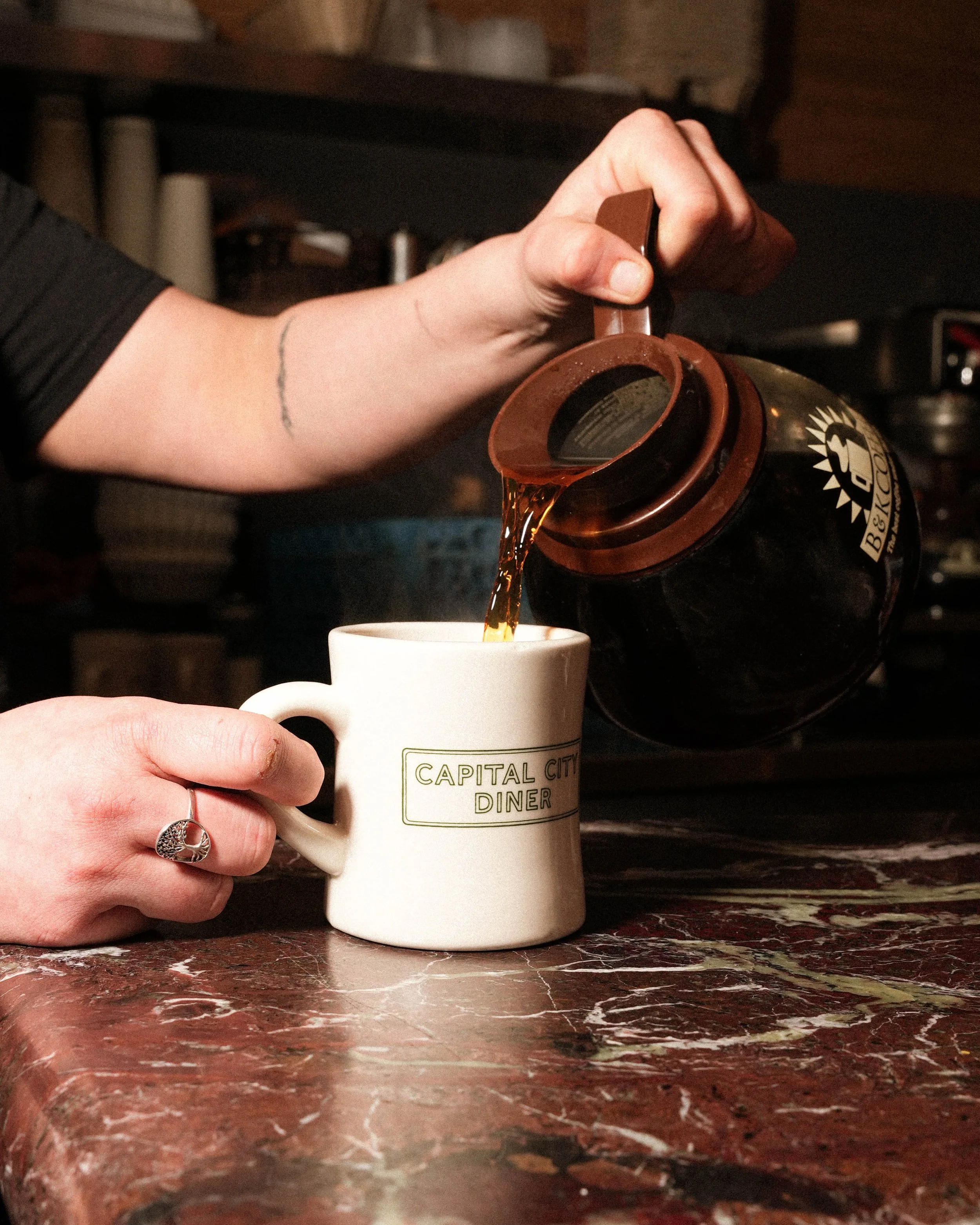 A person is pouring coffee from a thermos into a white mug with the words 'Capital City Diner' written on it, sitting on a marble countertop.