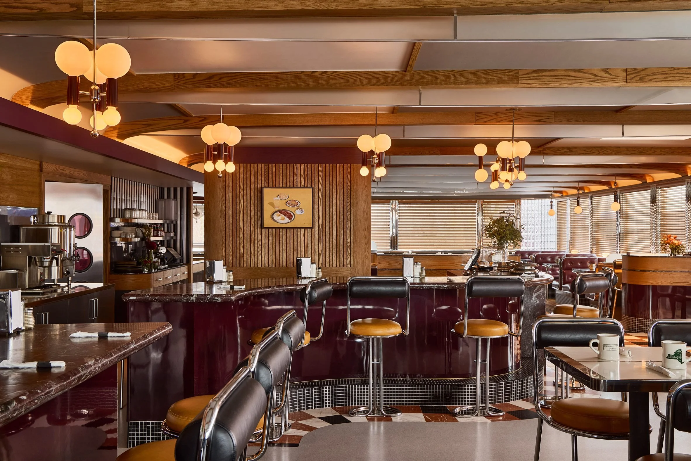 Interior of a retro-style diner with multiple hanging globe light fixtures, wooden wall paneling, and large windows with blinds. The counter has black chairs with mustard-colored cushions, and there are tables with cups and utensils. A framed artwork