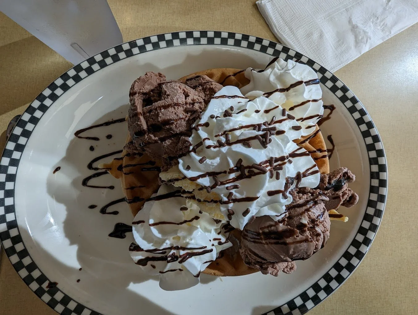 Chocolate ice cream, whipped cream, chocolate syrup, waffle, and sprinkles on a checkered plate.