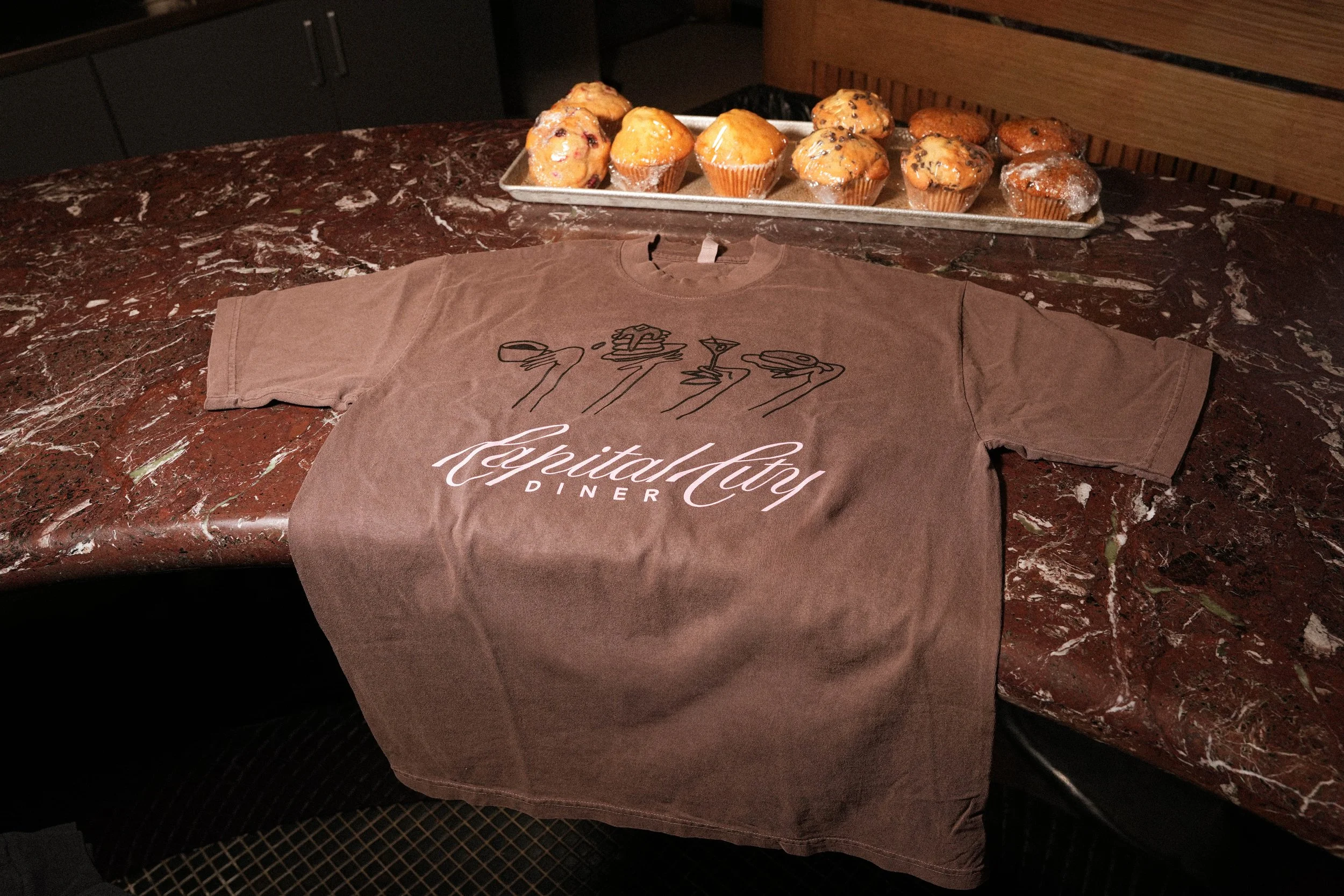 A brown T-shirt with the words 'Capital City Diner' and a line art logo of a hand holding a drink is laid out on a reddish marble table. Behind it, a tray holds various muffins, some with chocolate chips and others without.