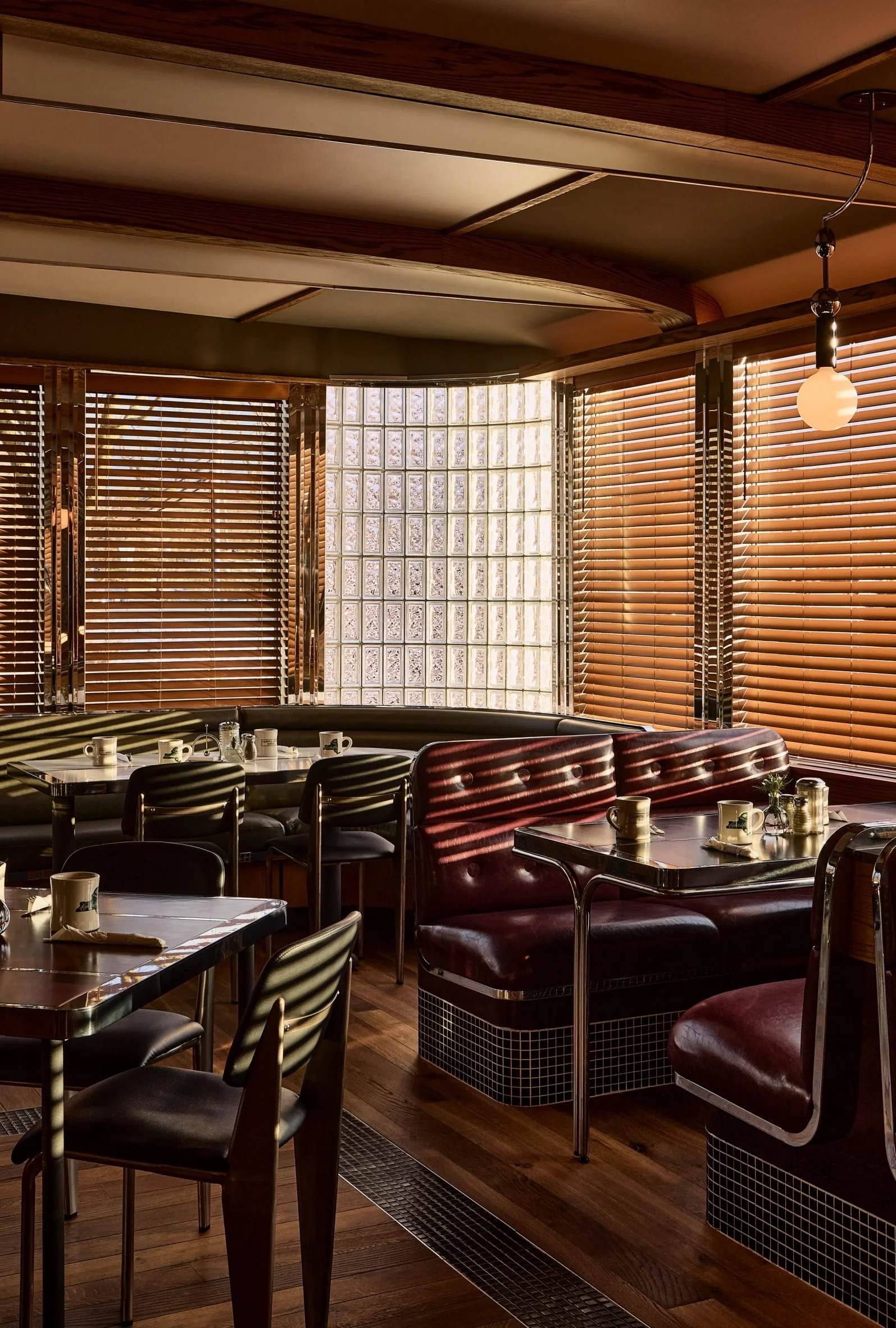 Dining area with wooden blinds, tables with cups and saucers, and vintage-style red leather booths.