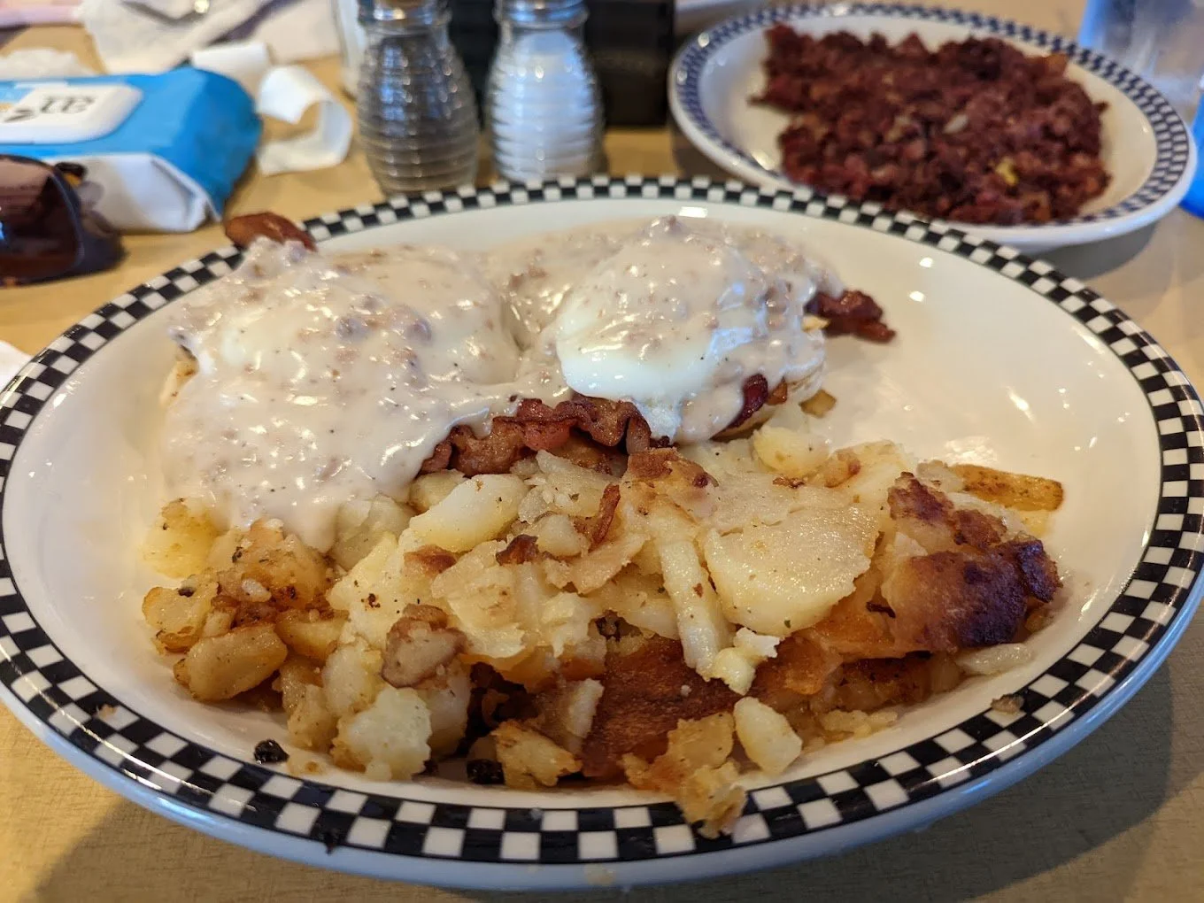 A breakfast plate with biscuits and gravy topped with two poached eggs, served with a side of breakfast potatoes. In the background, there is a bowl of hash and salt and pepper shakers on a wooden table.