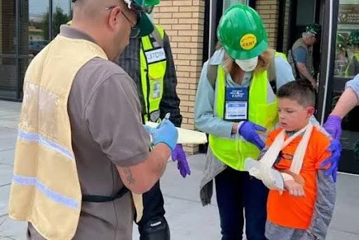 Construction workers and a young boy wearing safety gear and gloves, with one of the workers gently helping the boy put on a hard hat.