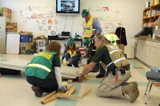 Four people in construction safety vests and helmets working on a project inside a room, possibly constructing or repairing something.