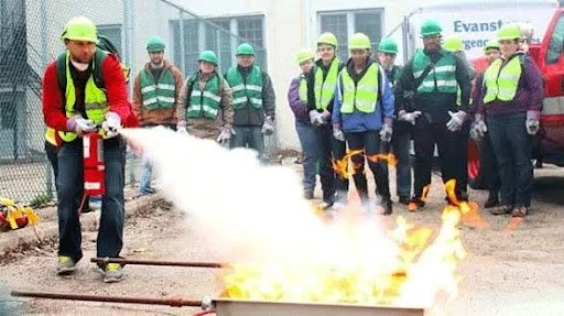 Fire safety training with an instructor wearing safety gear demonstrating the use of a fire extinguisher to a group of people in safety vests and helmets.