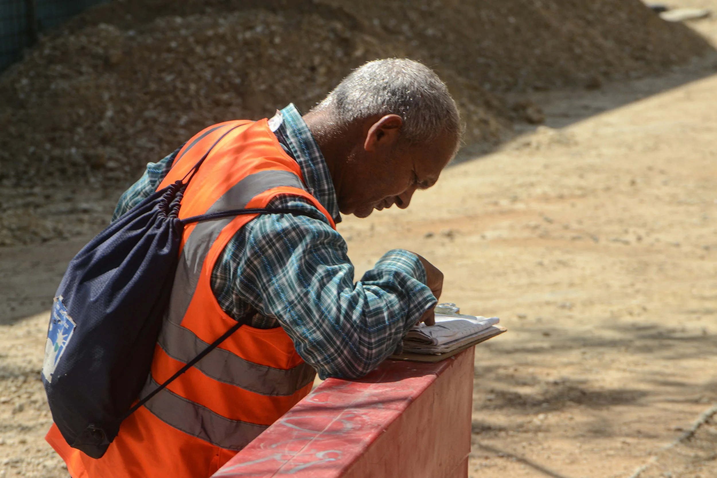 Historian or construction worker wearing an orange safety vest, leaning on a red barrier and examining documents outdoors with a pile of gravel in the background.