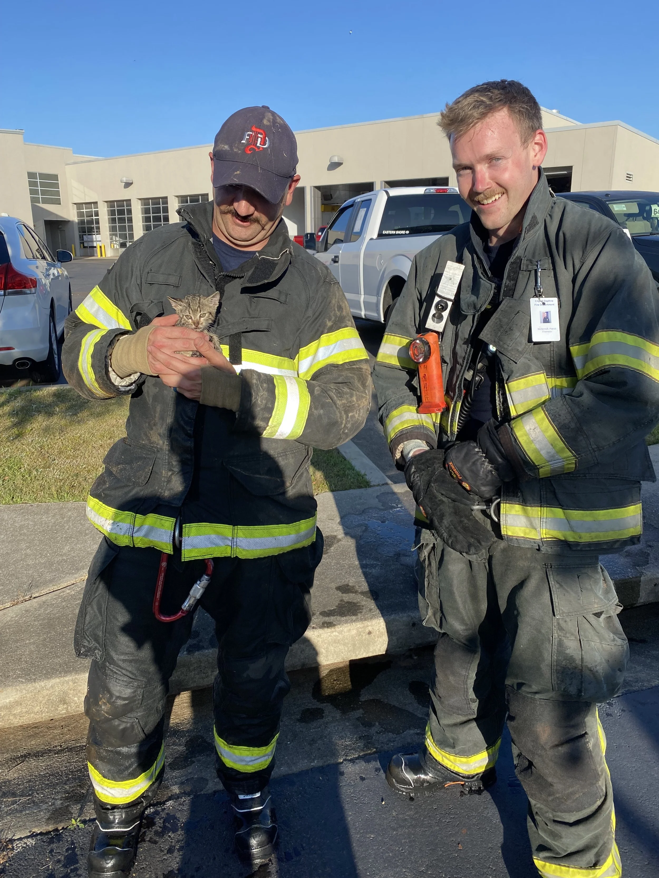 The Founder of Noah's Hammer and another firefighter holding a small kitten, standing outside in front of a parking lot on a sunny day.