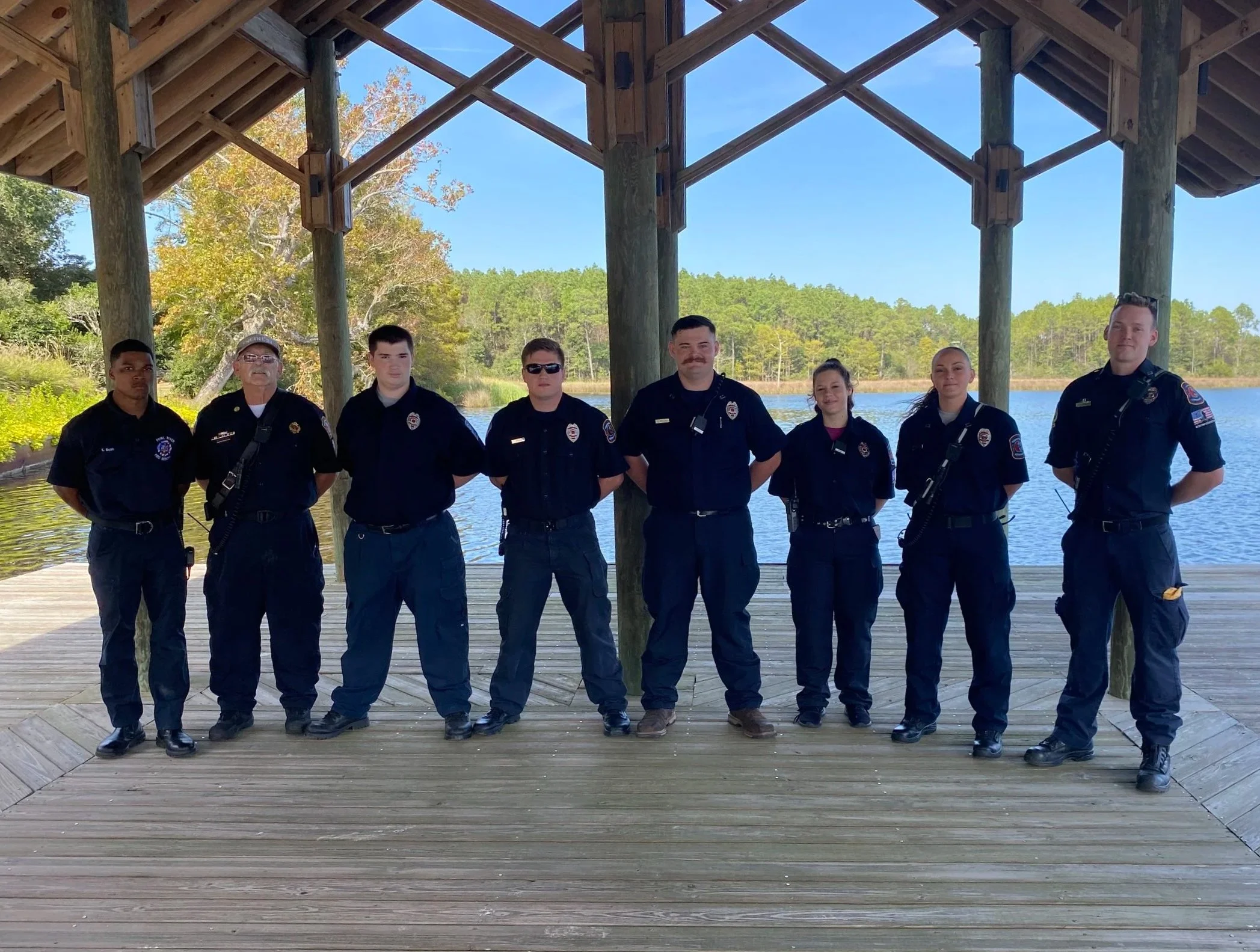 The Founder of Noah's Hammer and his volunteer fire department standing side by side on a wooden dock under a pavilion with a lake and trees in the background.