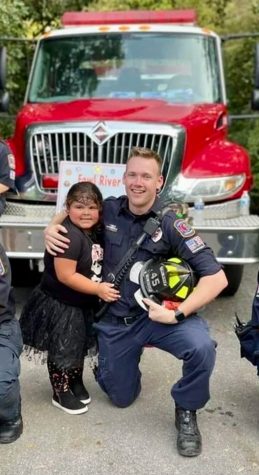 A young girl hugging the founder of Noah's Hammer, a firefighter, in front of a red fire truck at a halloween event.