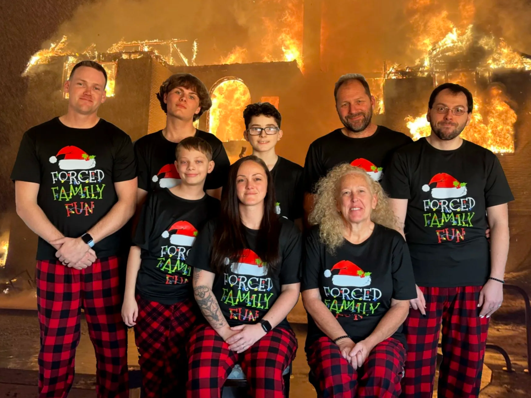 The Founder of Noah's Hammer and his family posing for a Christmas photo. They are dressed in matching black T-shirts with Christmas hats and the words 'Forced Family Fun' printed on them, posing in front of a large house fire background.