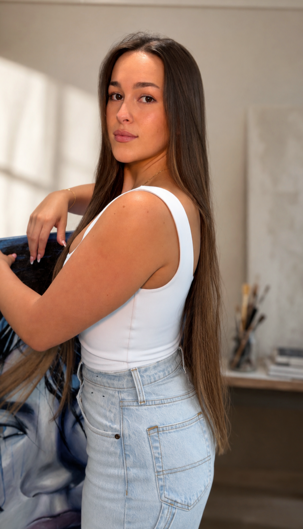 A young woman with long brown hair and light skin, wearing a white sleeveless top and light denim jeans, standing indoors and looking at the camera.